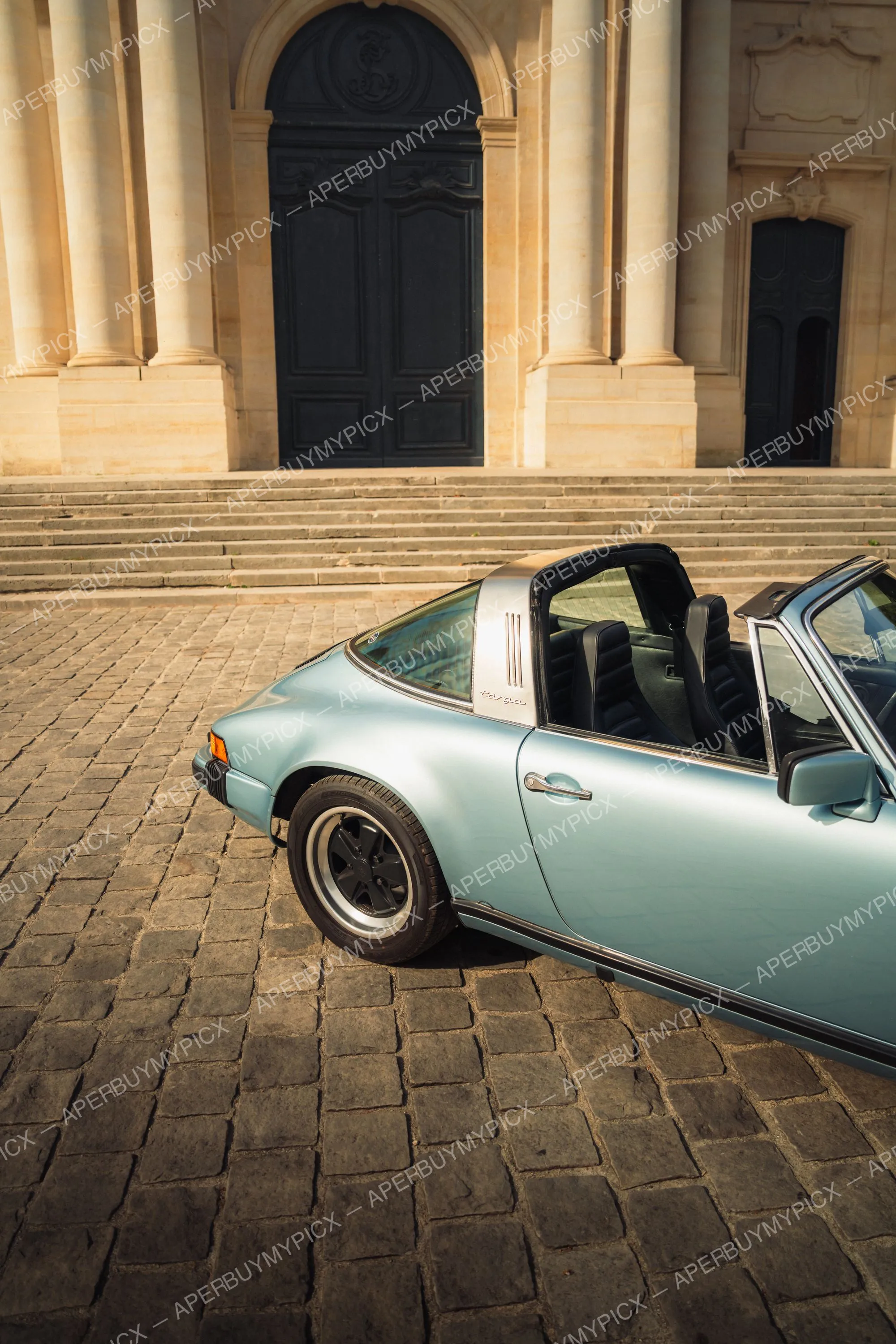Porsche 911 SC Targa bleu clair décapotée garée sur des pavés devant un bâtiment classique avec colonnes et grandes portes noires