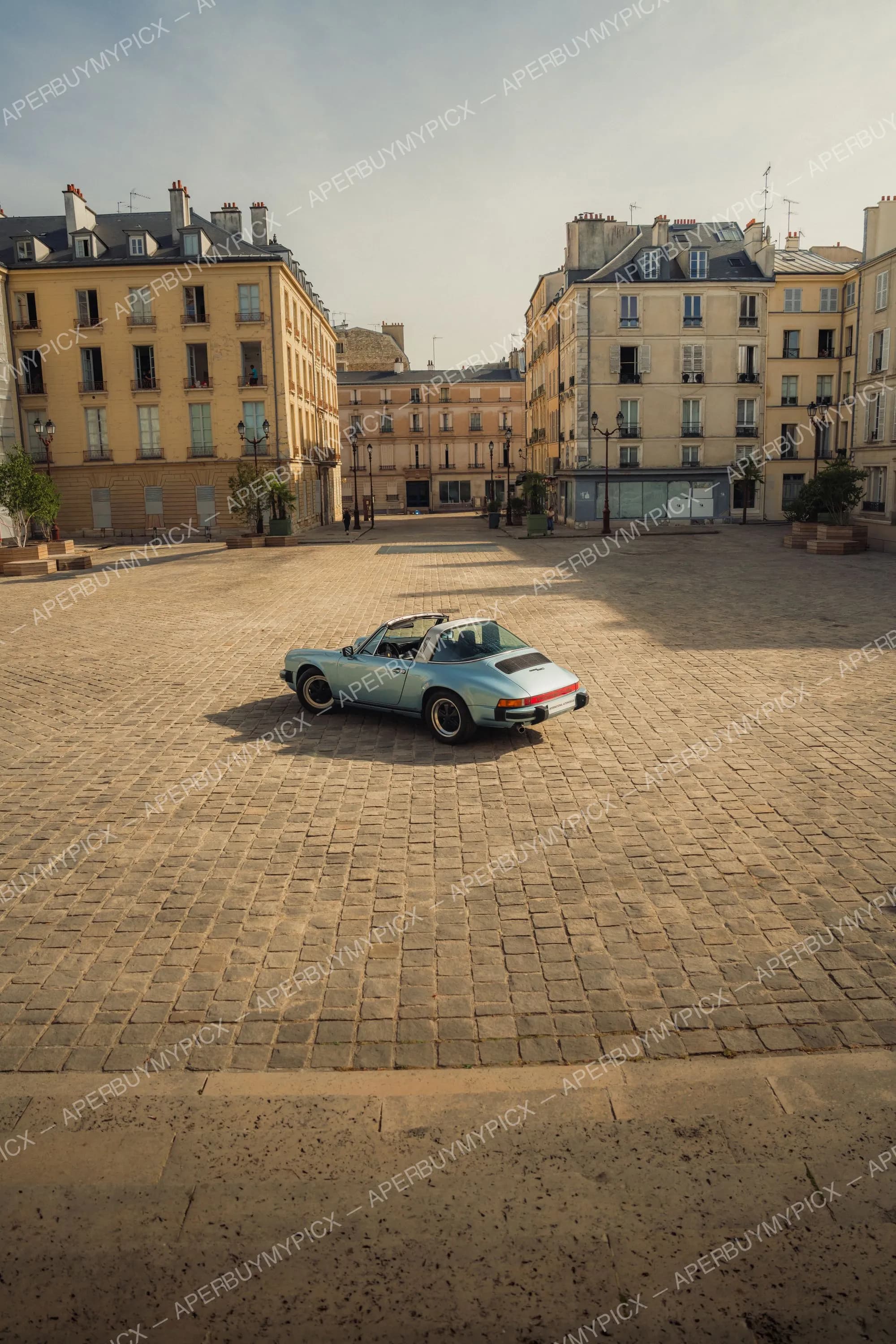 Porsche 911 SC Targa bleu clair garée sur une vaste place pavée bordée d'immeubles haussmanniens parisiens sous une lumière dorée.