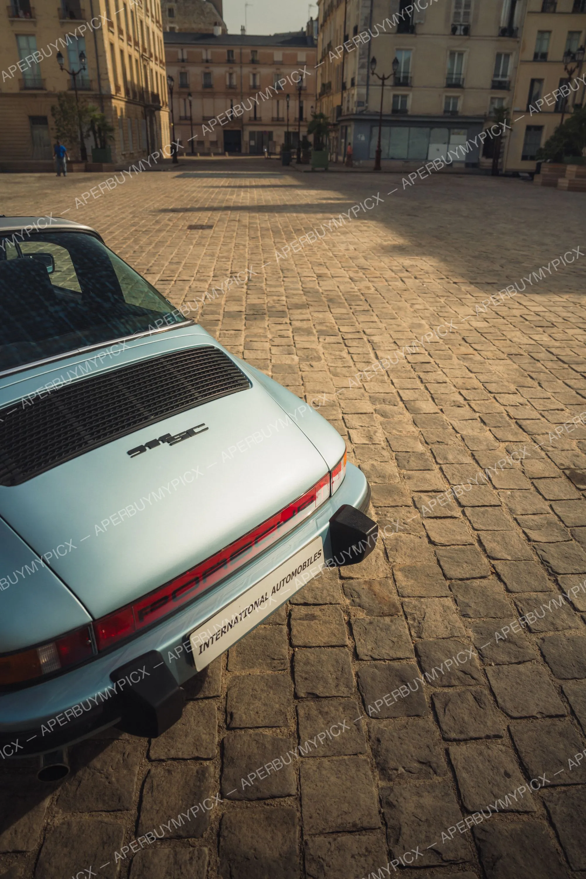 Arrière d'une Porsche 911 SC Targa bleu clair vintage garée sur une place pavée baignée de lumière dorée à Paris, avec immeubles haussmanniens en arrière-plan.