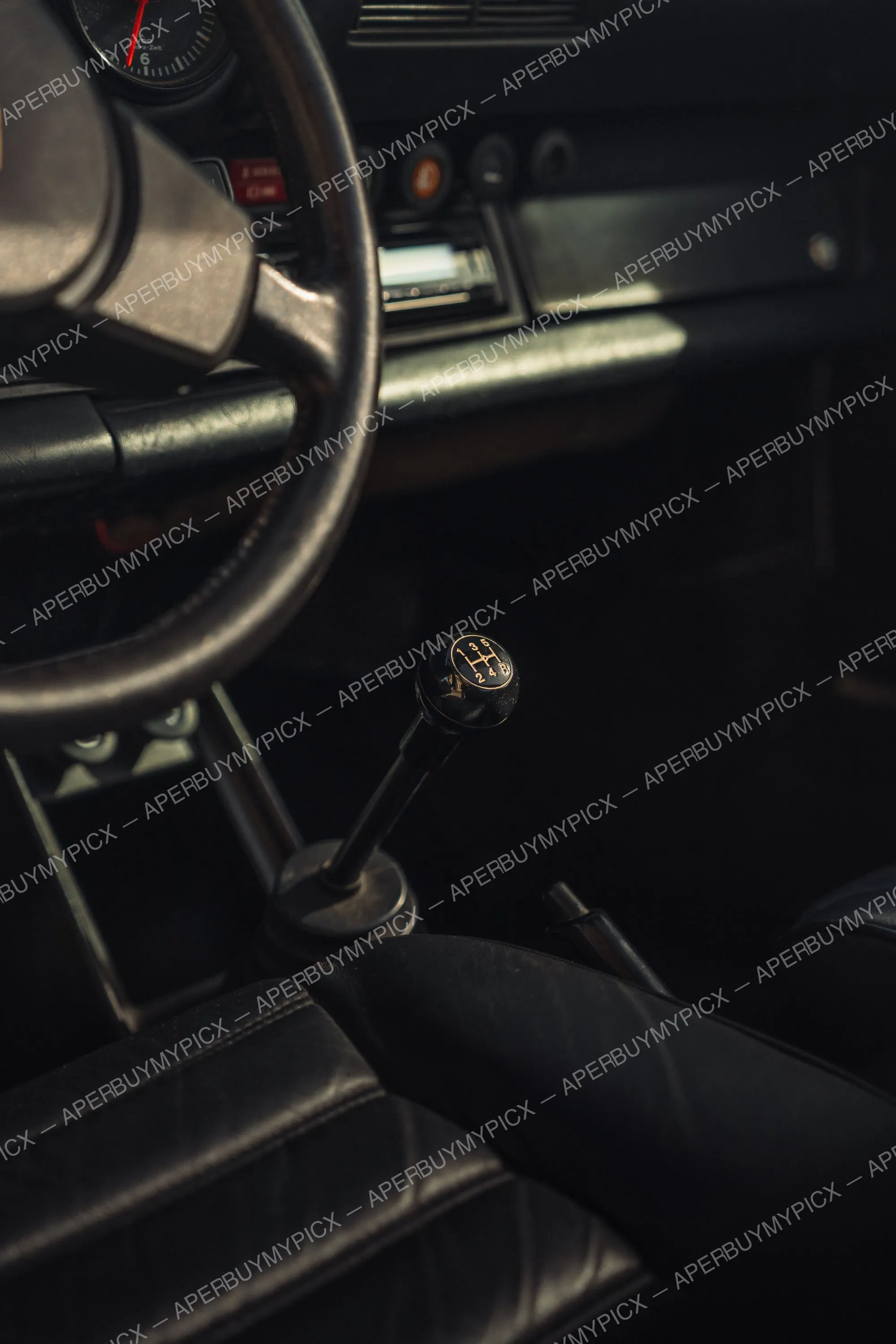 Gros plan sur le pommeau de levier de vitesse à 5 rapports d'une Porsche 911 SC Targa, avec le volant, le tableau de bord et les sièges en cuir noir en arrière-plan sombre