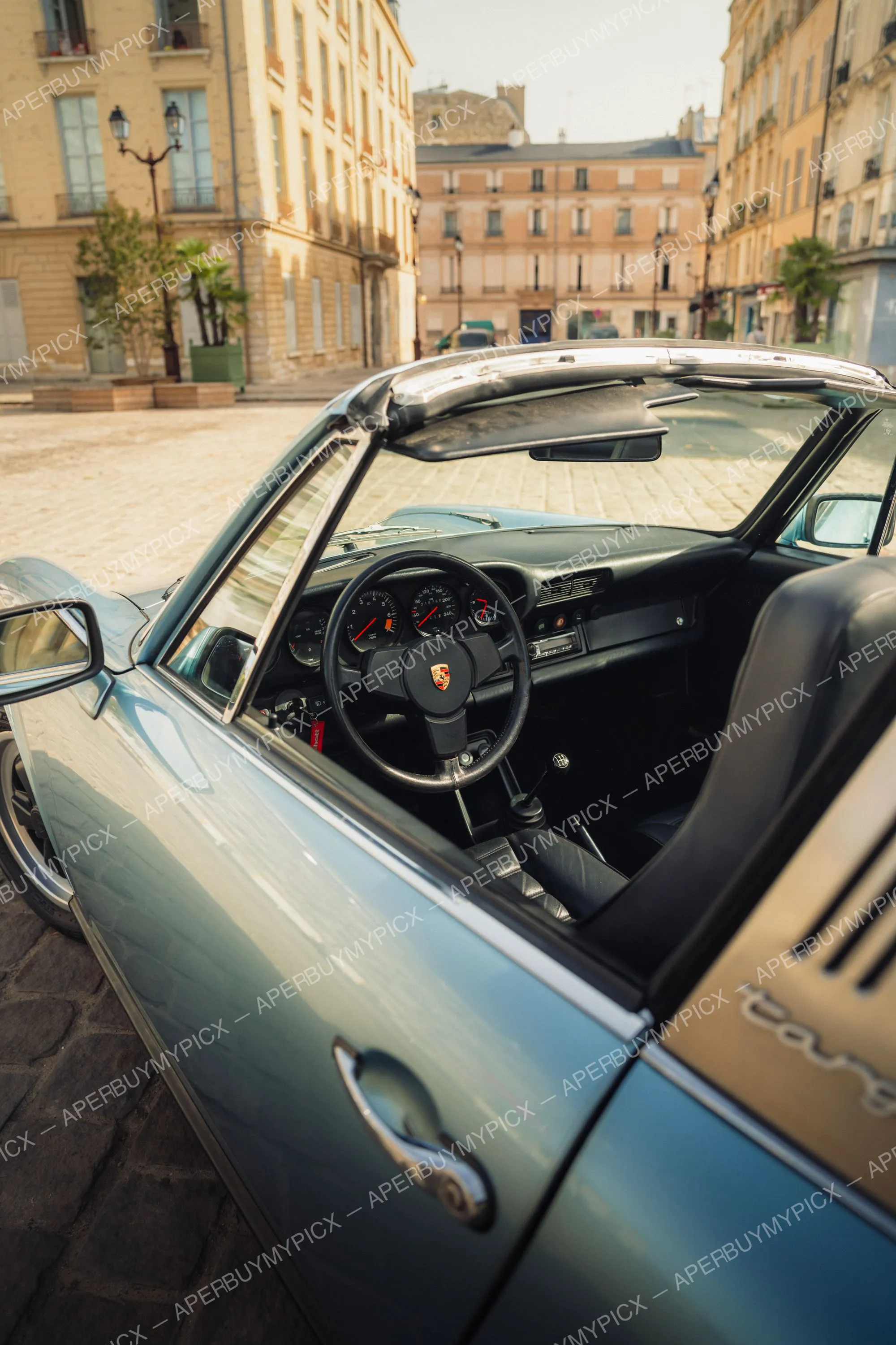 Porsche 911 SC Targa bleu clair vue depuis la vitre ouverte montrant le tableau de bord, le volant à écusson Porsche et l'intérieur en cuir noir, garée sur une place pavée parisienne