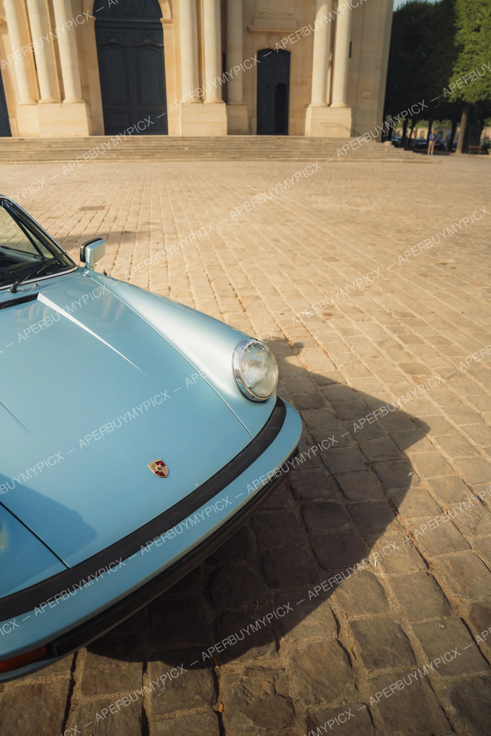 Avant d'une Porsche 911 SC Targa bleu clair vintage avec phare rond et écusson Porsche, garée sur des pavés devant un bâtiment classique à colonnes en pierre au soleil couchant.