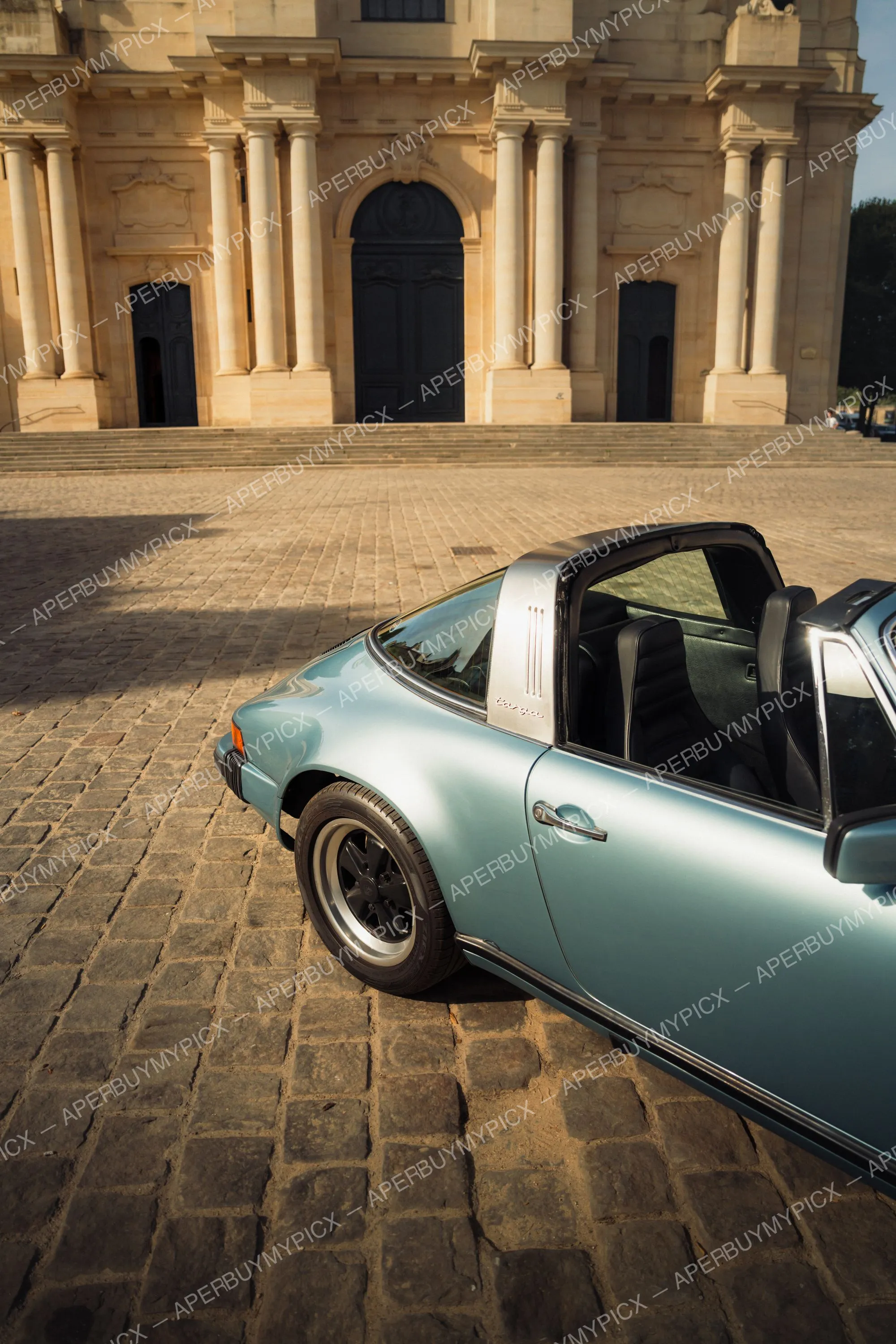 Porsche 911 SC Targa bleu métallisé garée sur une place pavée devant une imposante façade baroque en pierre avec colonnes et grande porte sombre au coucher du soleil