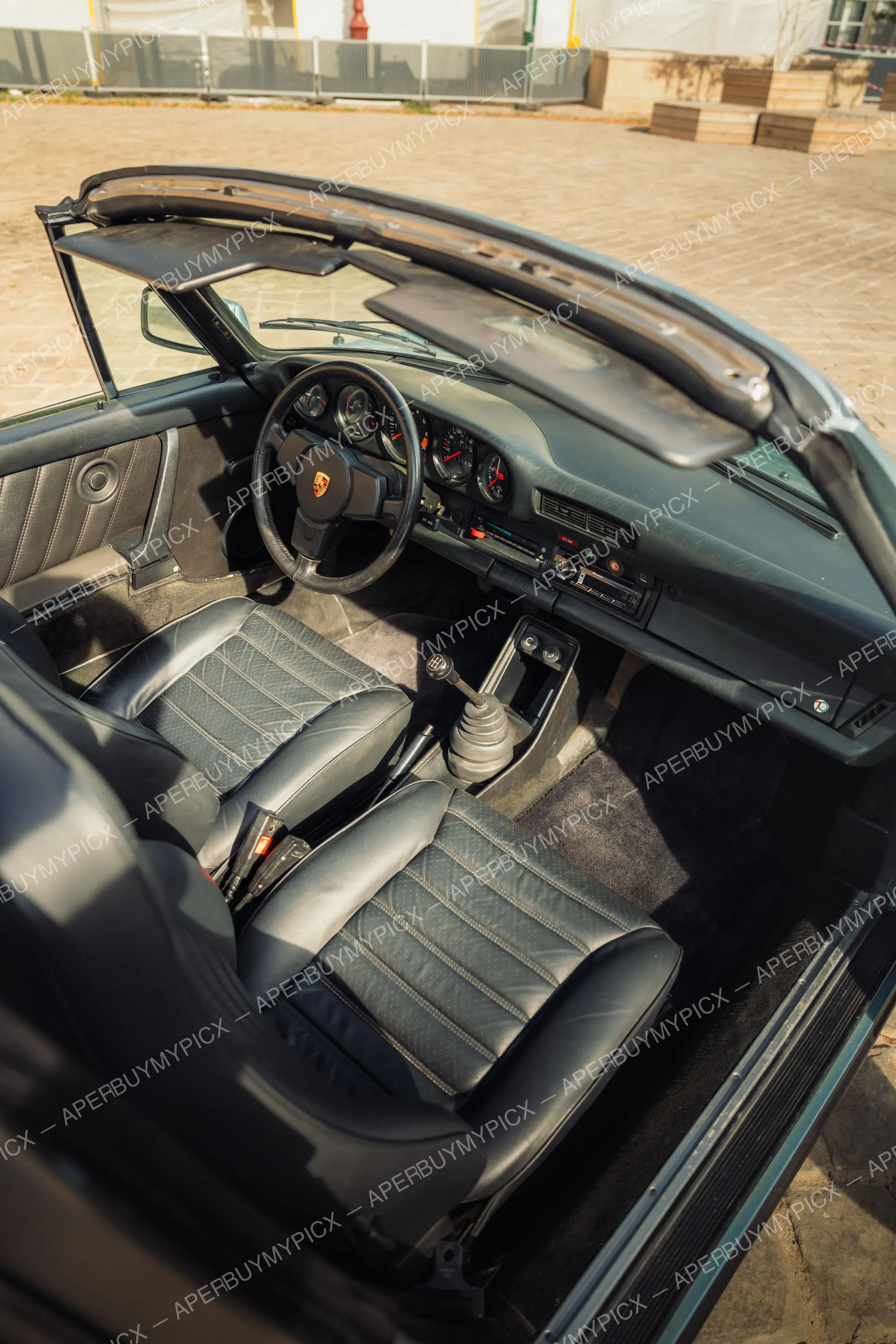 Intérieur d'une Porsche 911 SC Targa vue de dessus avec toit retiré, sièges en cuir noir matelassé, volant Porsche, levier de vitesses manuel et tableau de bord à cadrans classiques.