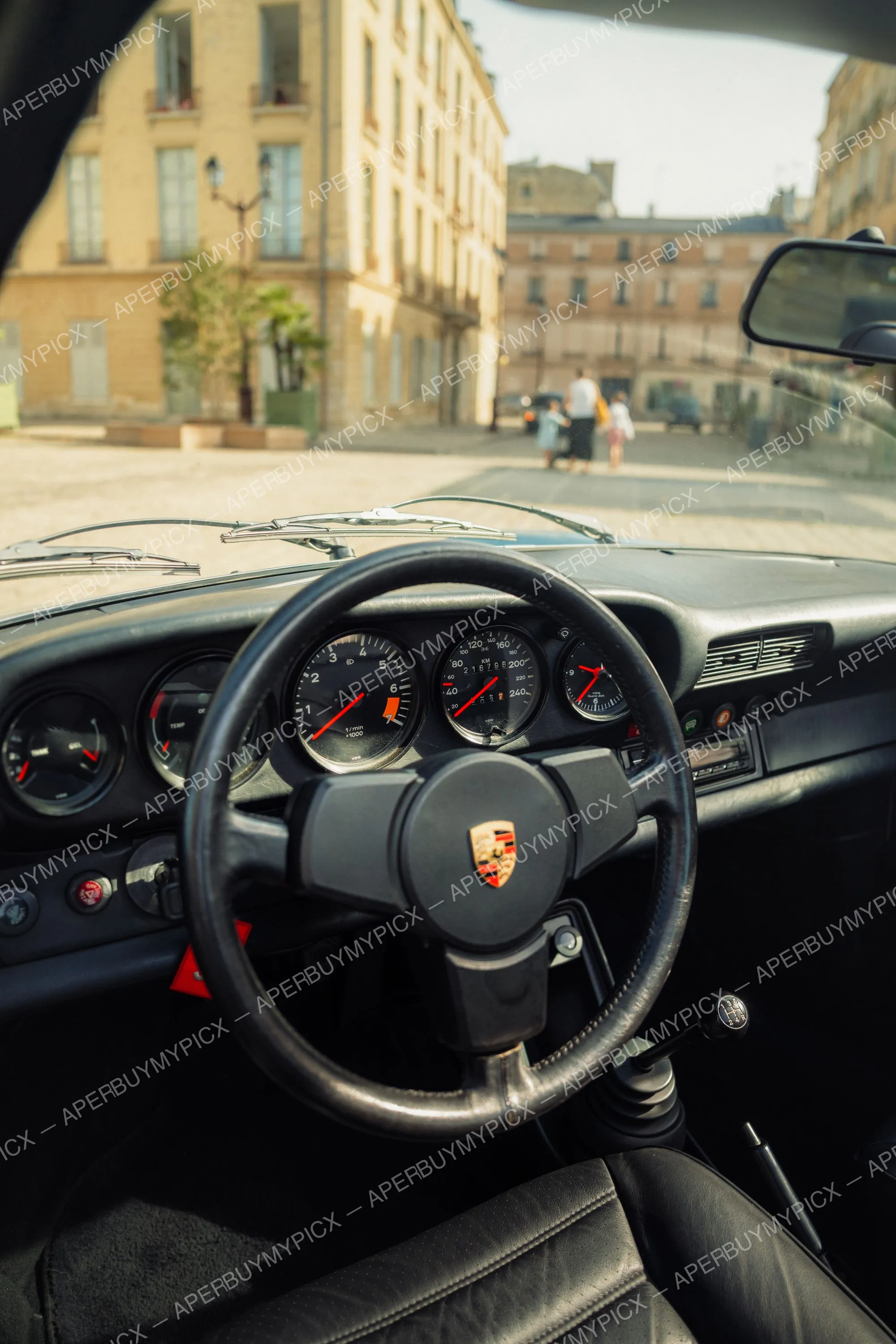 Vue de l'intérieur d'une Porsche 911 SC classique avec volant en cuir, logo Porsche, compteurs et levier de vitesse manuel, à travers le pare-brise une rue parisienne ensoleillée avec bâtiments haussmanniens et piétons.