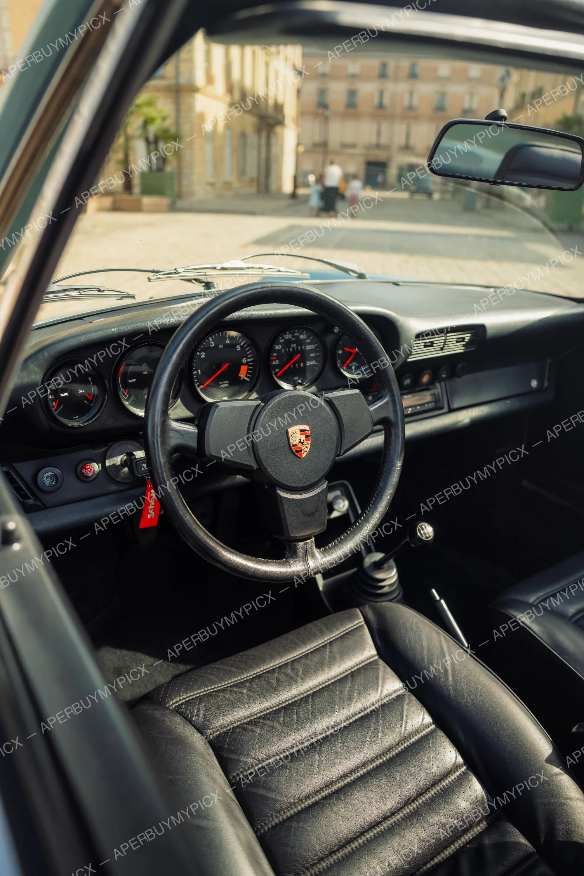 Intérieur d'une Porsche 911 SC Targa avec volant noir à logo Porsche, tableau de bord à compteurs analogiques, siège en cuir noir capitonné et vue sur une place urbaine à travers le pare-brise
