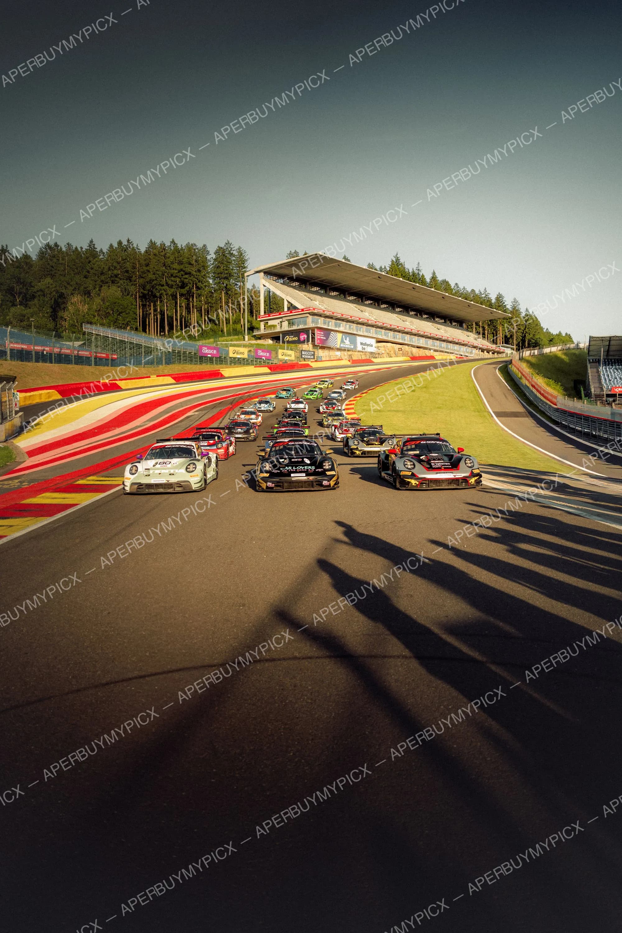 Peloton de voitures de course Porsche au départ sur le circuit de Spa-Francorchamps, avec tribunes, vibreurs rouges et jaunes et forêt en arrière-plan sous un ciel dégagé.