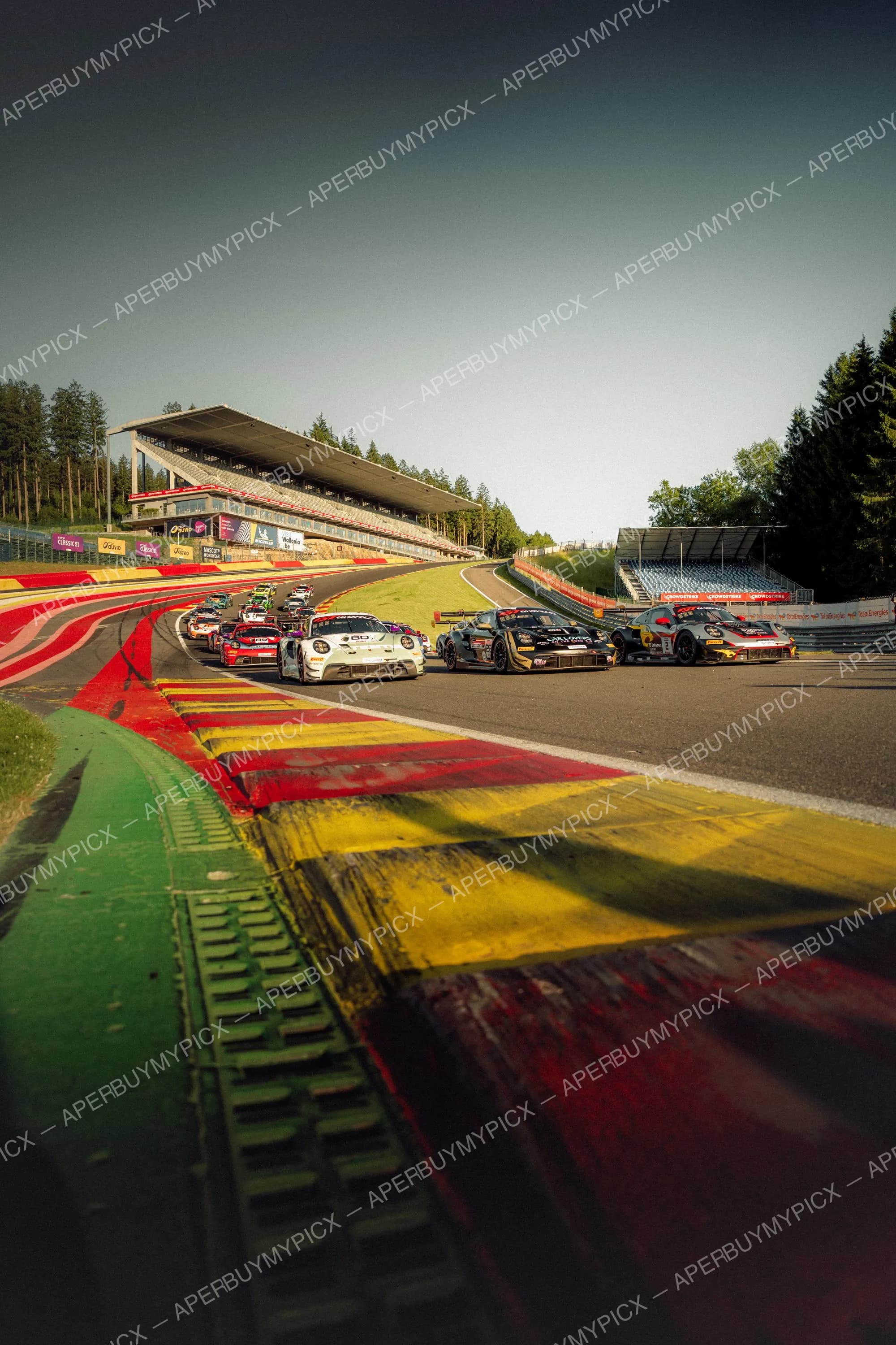 Peloton de Porsche 911 GT3 en pleine course sur le circuit de Spa-Francorchamps, franchissant les vibreurs rouges et jaunes avec les tribunes et la forêt en arrière-plan sous un ciel dégagé.