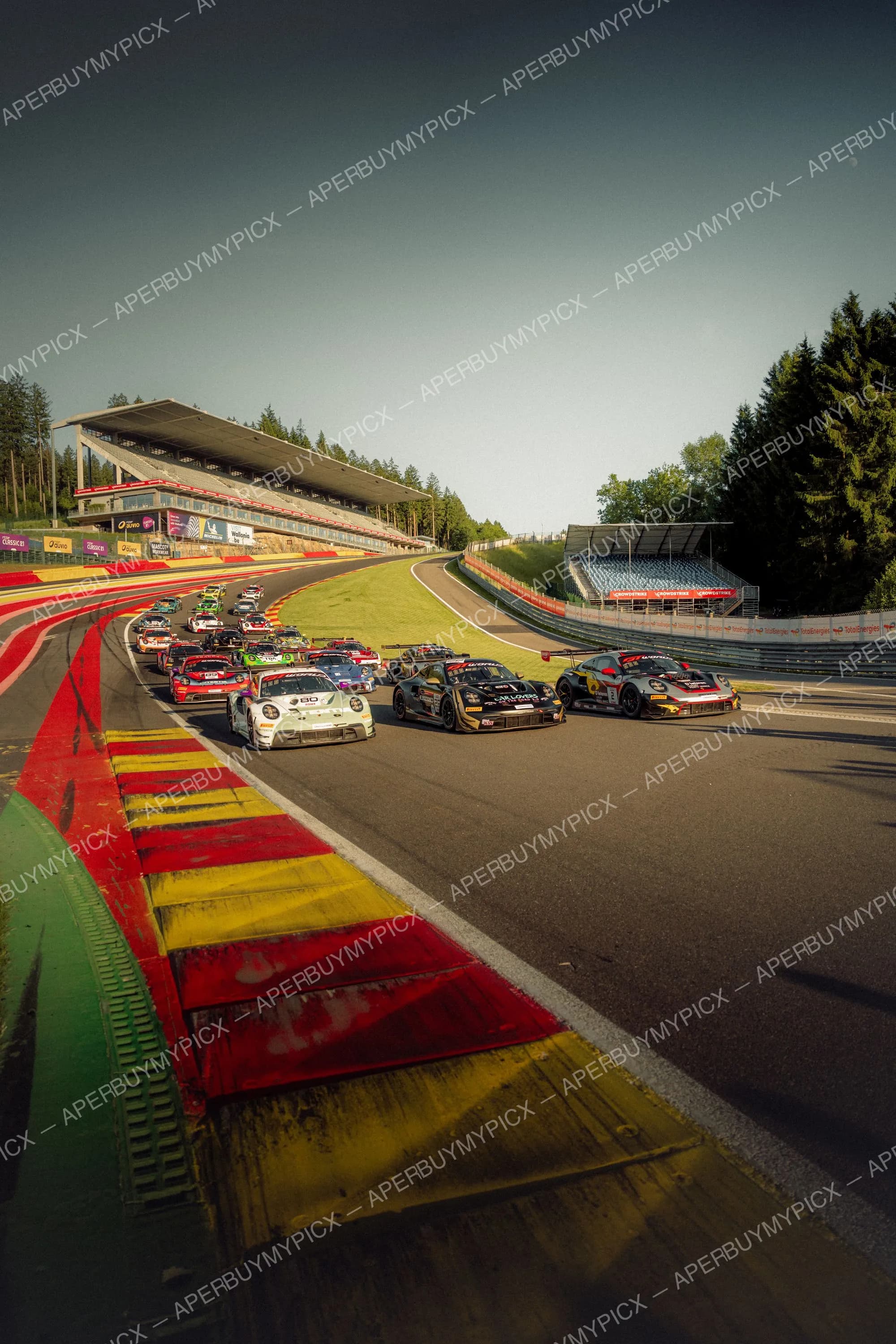 Peloton de voitures de course GT, principalement des Porsche 911, au départ sur le circuit de Spa-Francorchamps avec vibreurs rouge et jaune, tribunes et forêt en arrière-plan sous un ciel dégagé.