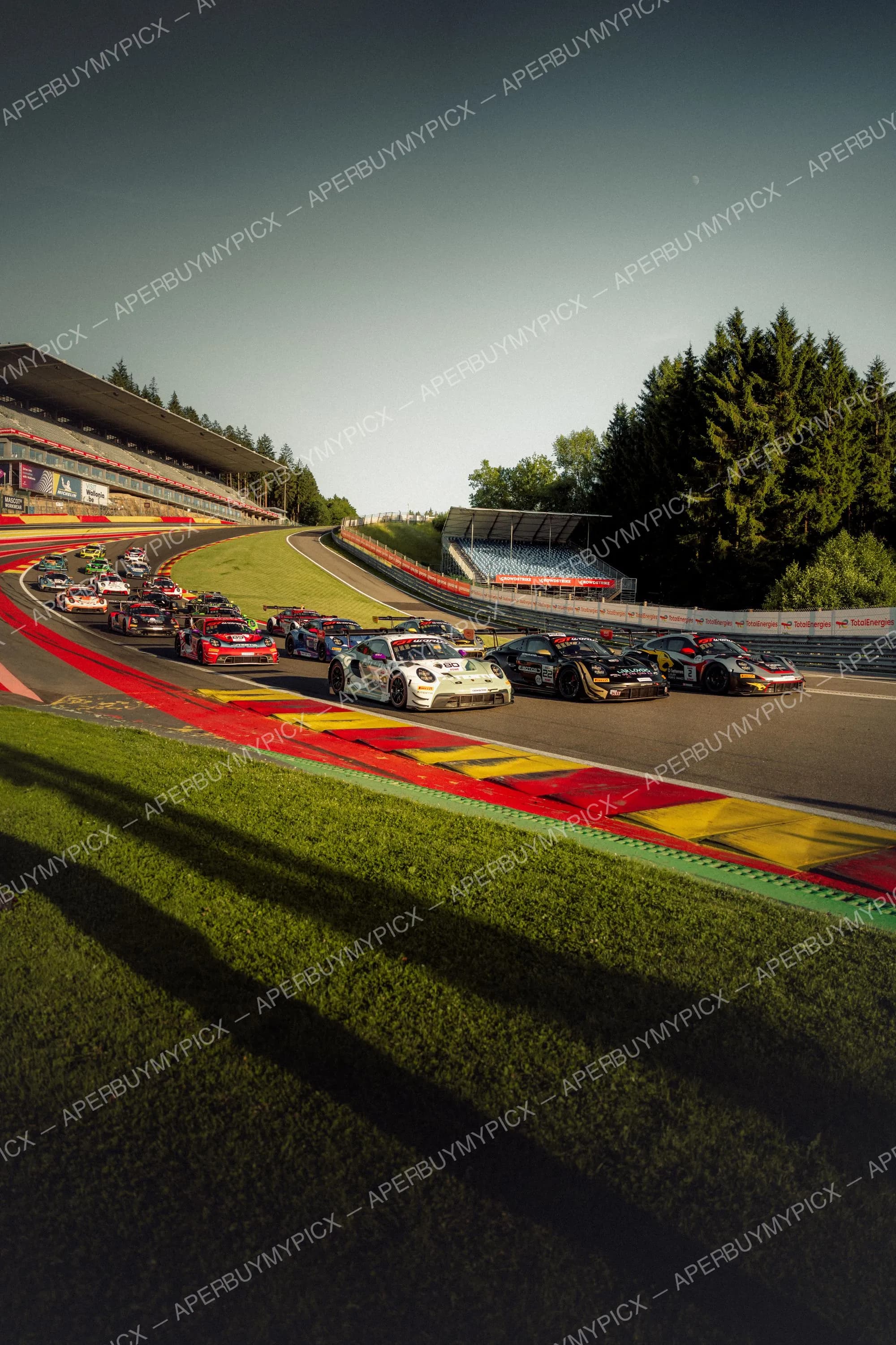 Peloton de voitures de course Porsche abordant un virage sur le circuit de Spa-Francorchamps, avec vibreurs rouges et jaunes, tribunes et forêt en arrière-plan sous une lumière de fin d'après-midi.