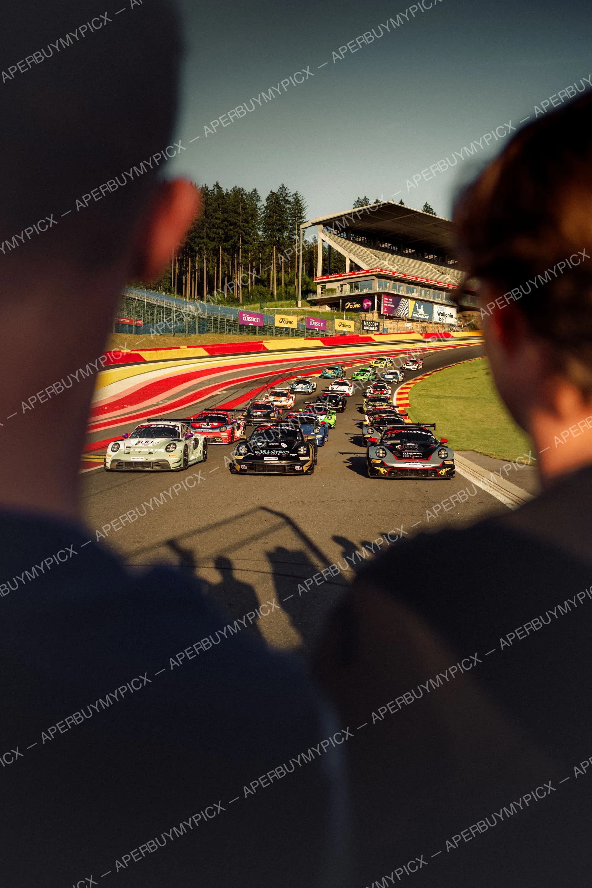 Peloton de voitures de course Porsche 911 au départ sur le circuit de Spa-Francorchamps, vu par-dessus l'épaule de spectateurs, avec vibreurs rouges et blancs, tribune et forêt en arrière-plan.