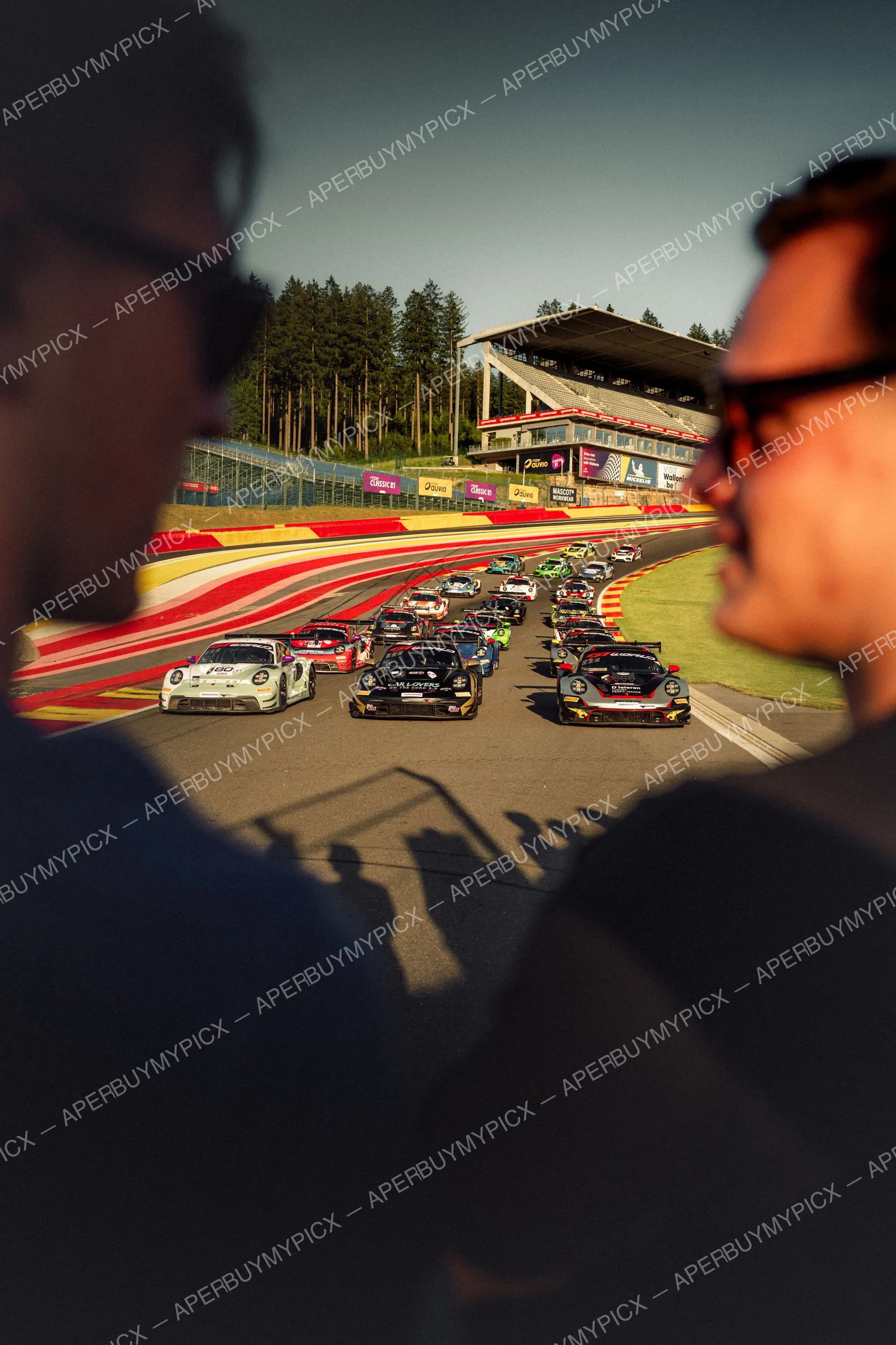 Peloton de voitures de course GT Porsche s'élançant sur le circuit de Spa-Francorchamps au coucher du soleil, vues à travers les silhouettes de deux spectateurs au premier plan, avec les tribunes et la forêt en arrière-plan.