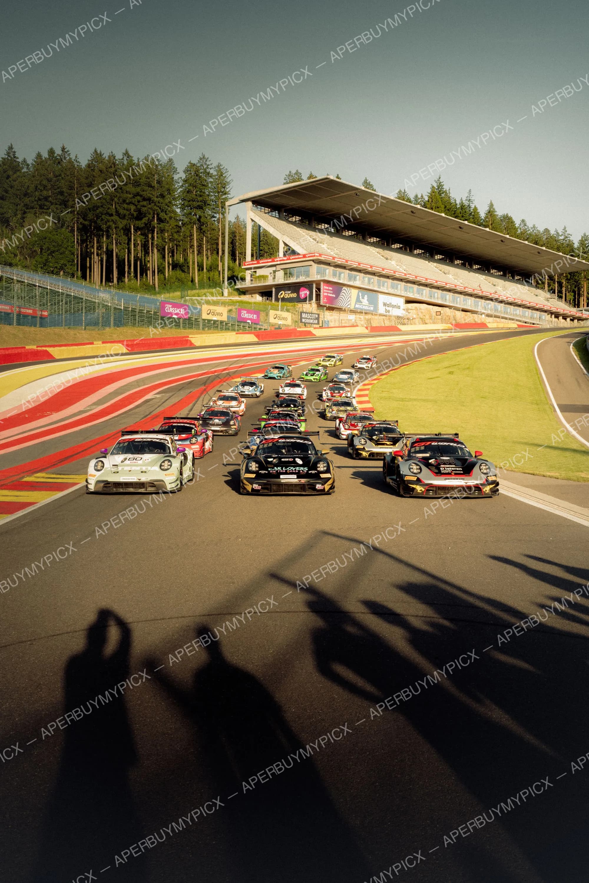 Grille de départ de voitures de course Porsche GT sur le circuit de Spa-Francorchamps, tribunes et forêt en arrière-plan sous un ciel dégagé.