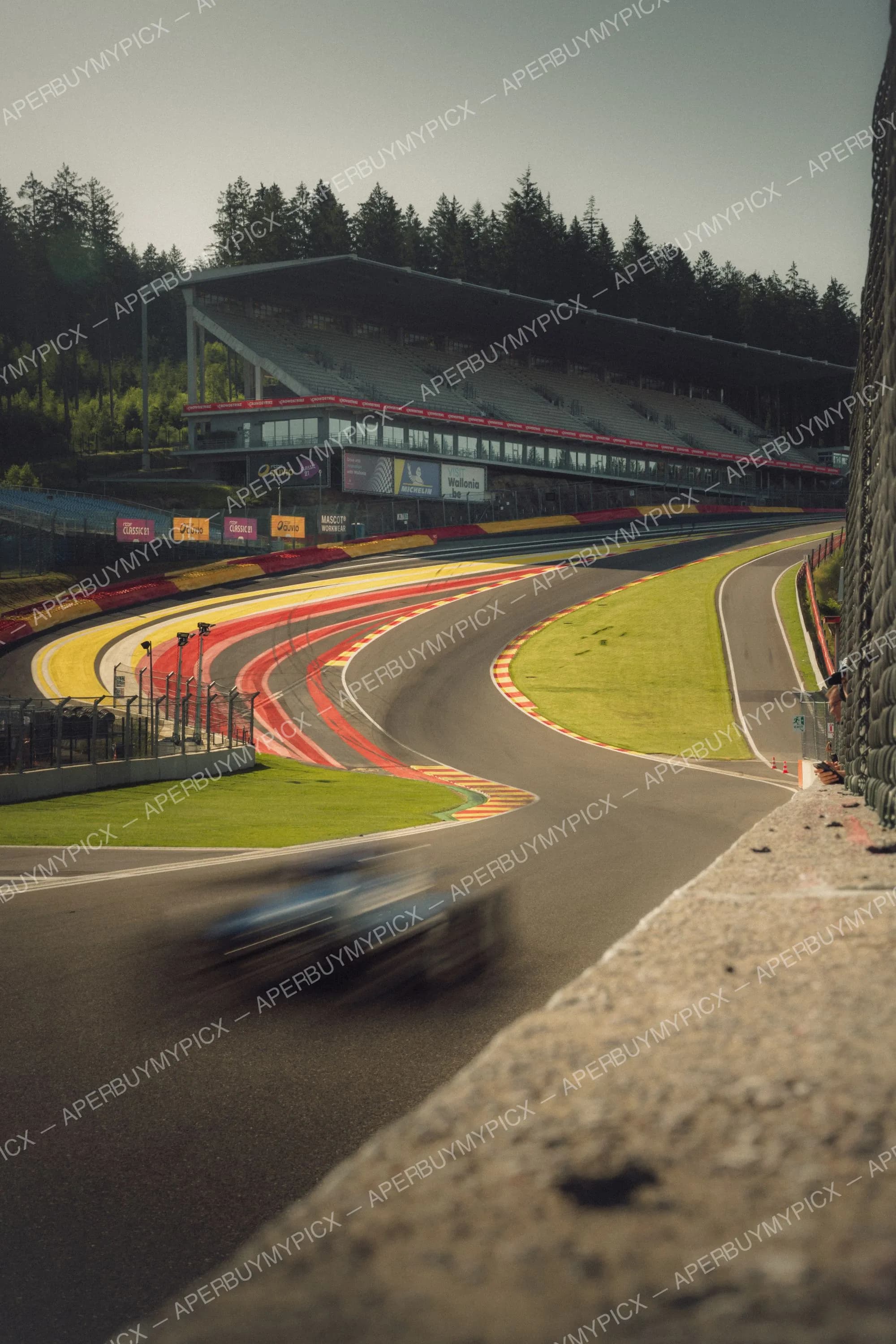 Voiture de course en mouvement flou sur le circuit de Spa-Francorchamps, vibreurs rouges et jaunes, tribune et forêt en arrière-plan au coucher du soleil