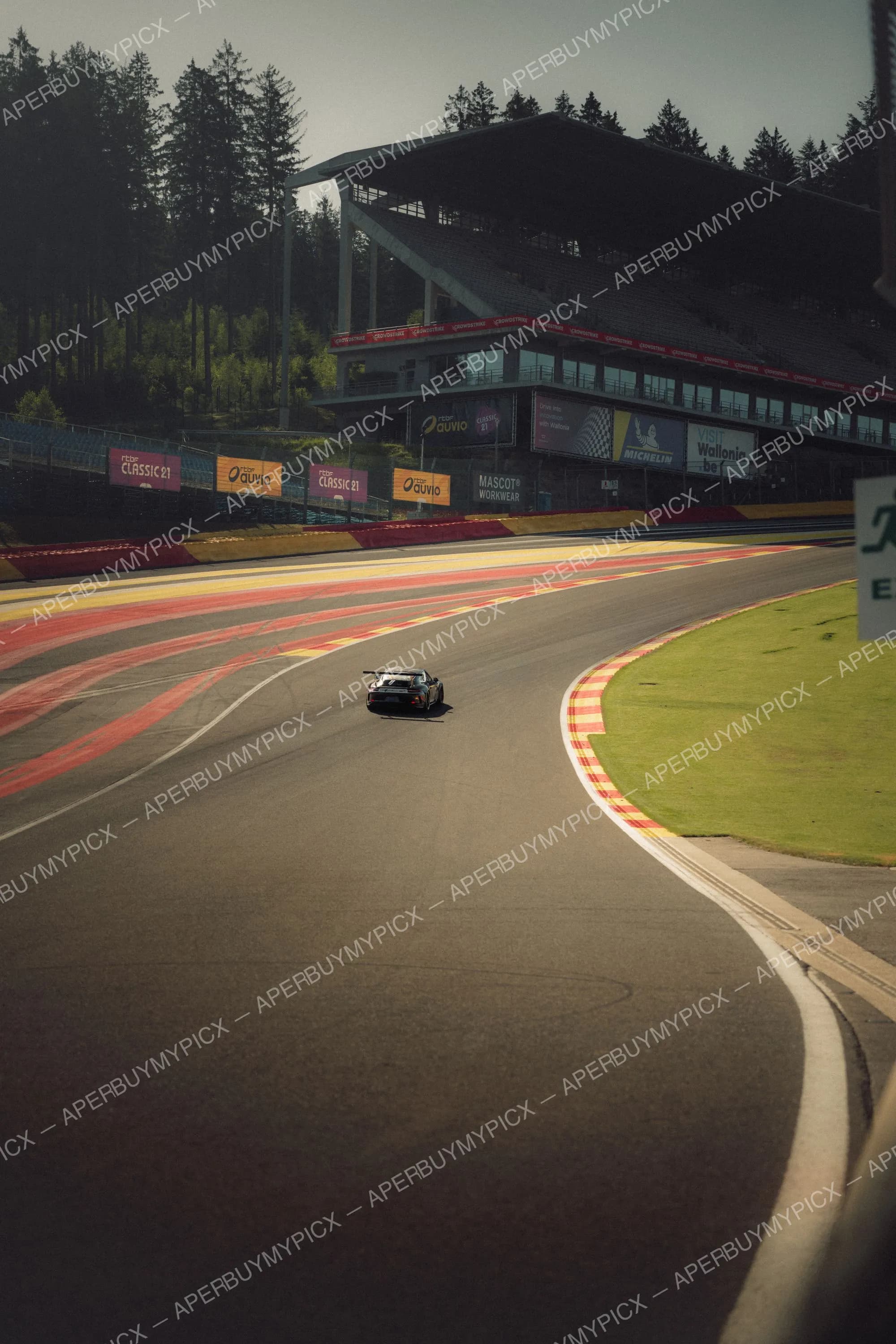Une Porsche GT3 négocie un virage sur le circuit de Spa-Francorchamps, avec les vibreurs rouge et jaune, la tribune et les panneaux publicitaires en arrière-plan sous une lumière rasante.