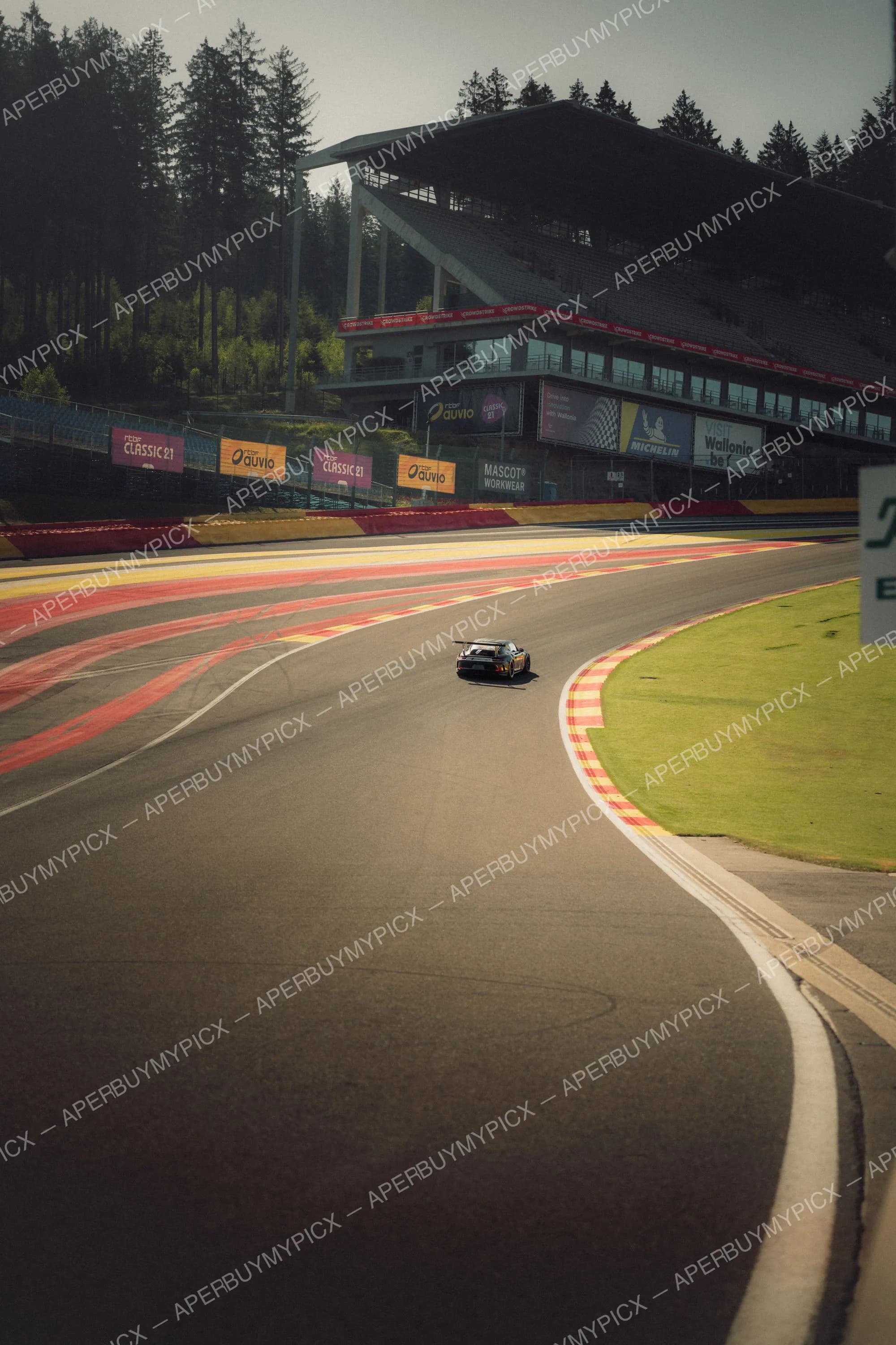 Une Porsche GT3 RS grise avec aileron arrière négocie un virage sur le circuit de Spa-Francorchamps, vibreurs rouges et jaunes, tribunes et panneaux publicitaires en arrière-plan.
