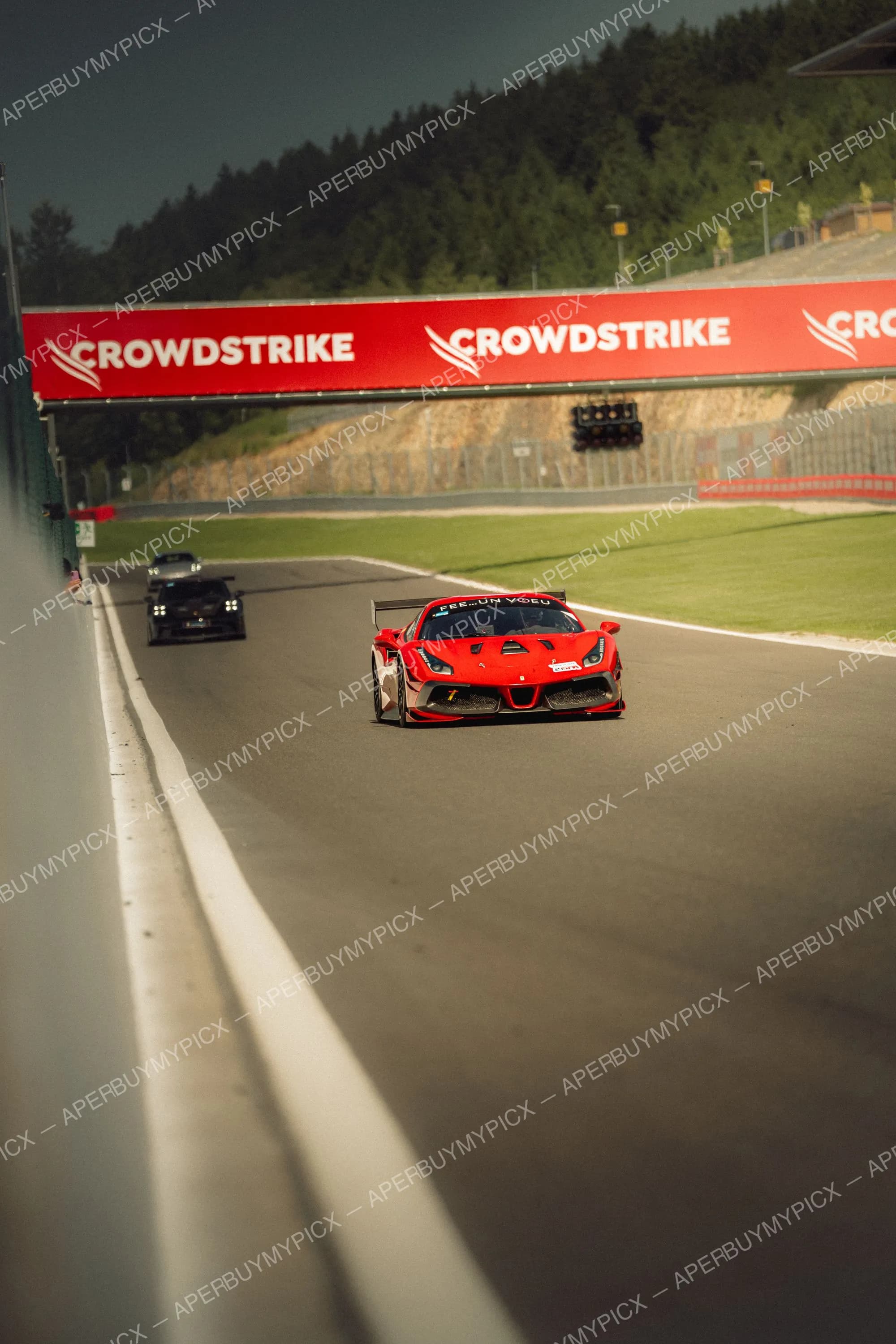 Ferrari 488 rouge de course sur le circuit de Spa-Francorchamps, suivie de voitures noires, sous un panneau publicitaire CrowdStrike rouge avec forêt en arrière-plan.