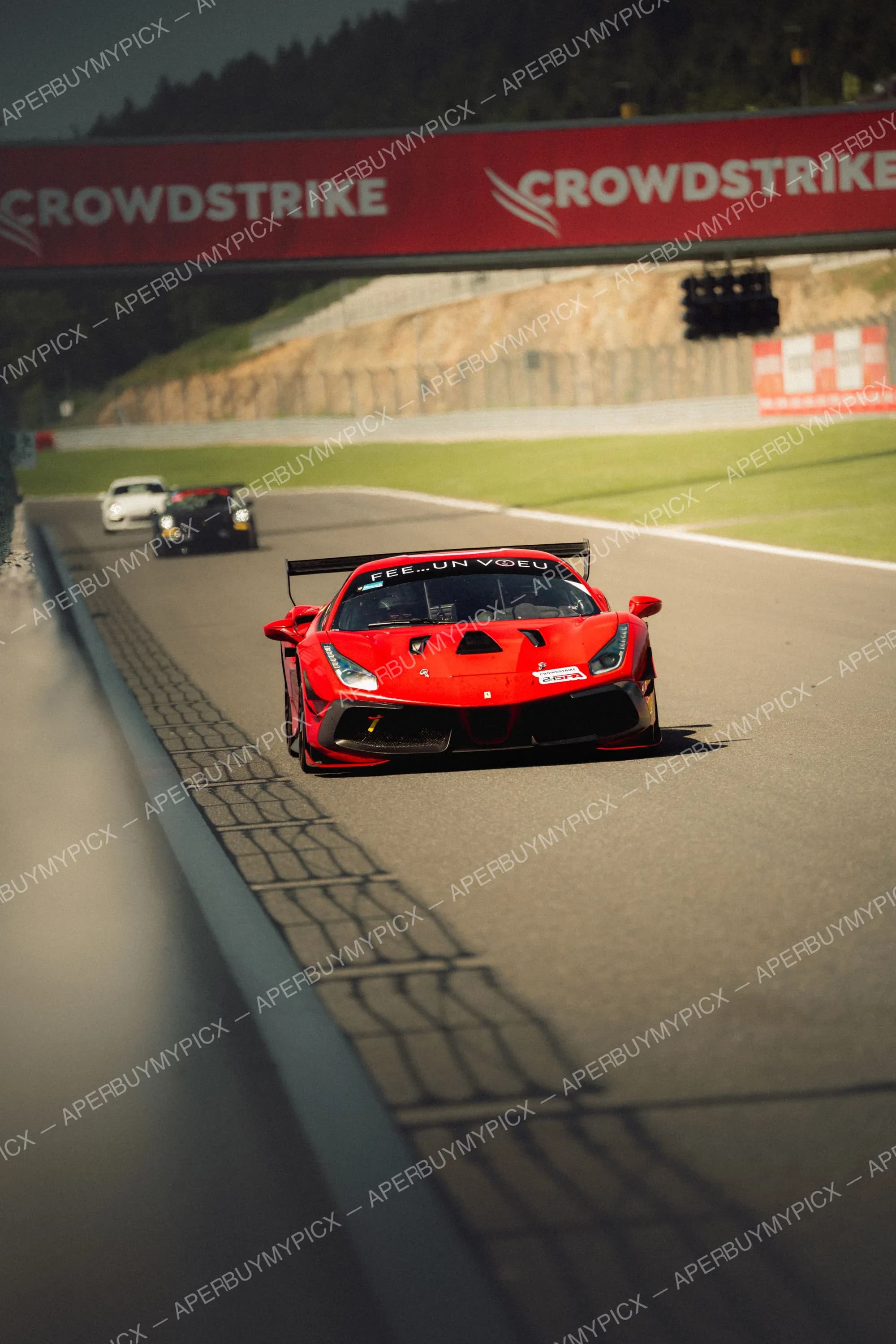 Ferrari 488 GT rouge portant l'inscription Fée...un vœu roulant sur le circuit de Spa-Francorchamps sous une passerelle CrowdStrike, suivie de deux voitures de course.