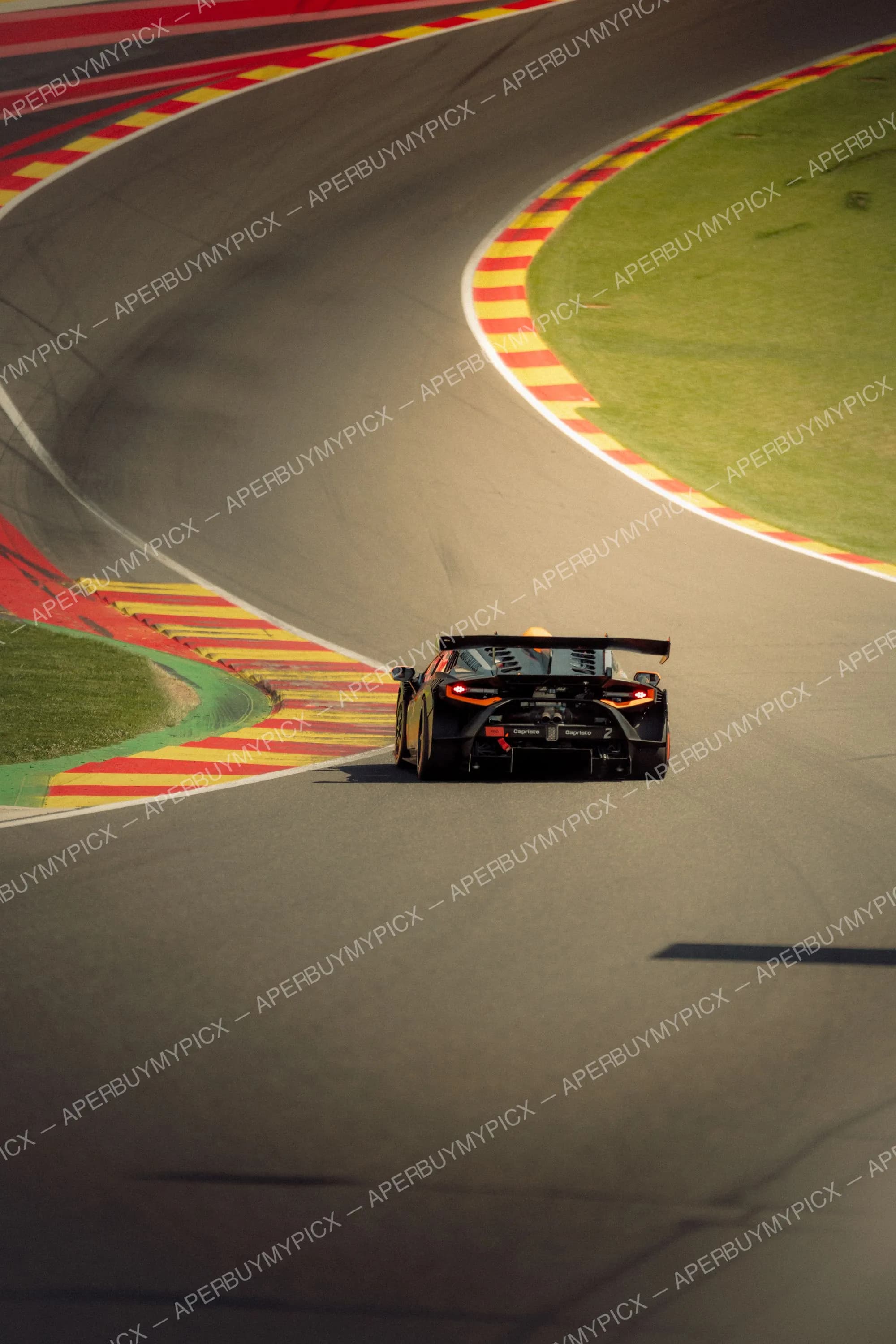 Voiture de course GT noire et orange abordant un virage sur le circuit de Spa-Francorchamps, avec vibreurs rouges et jaunes et pelouse verte en bordure de piste.