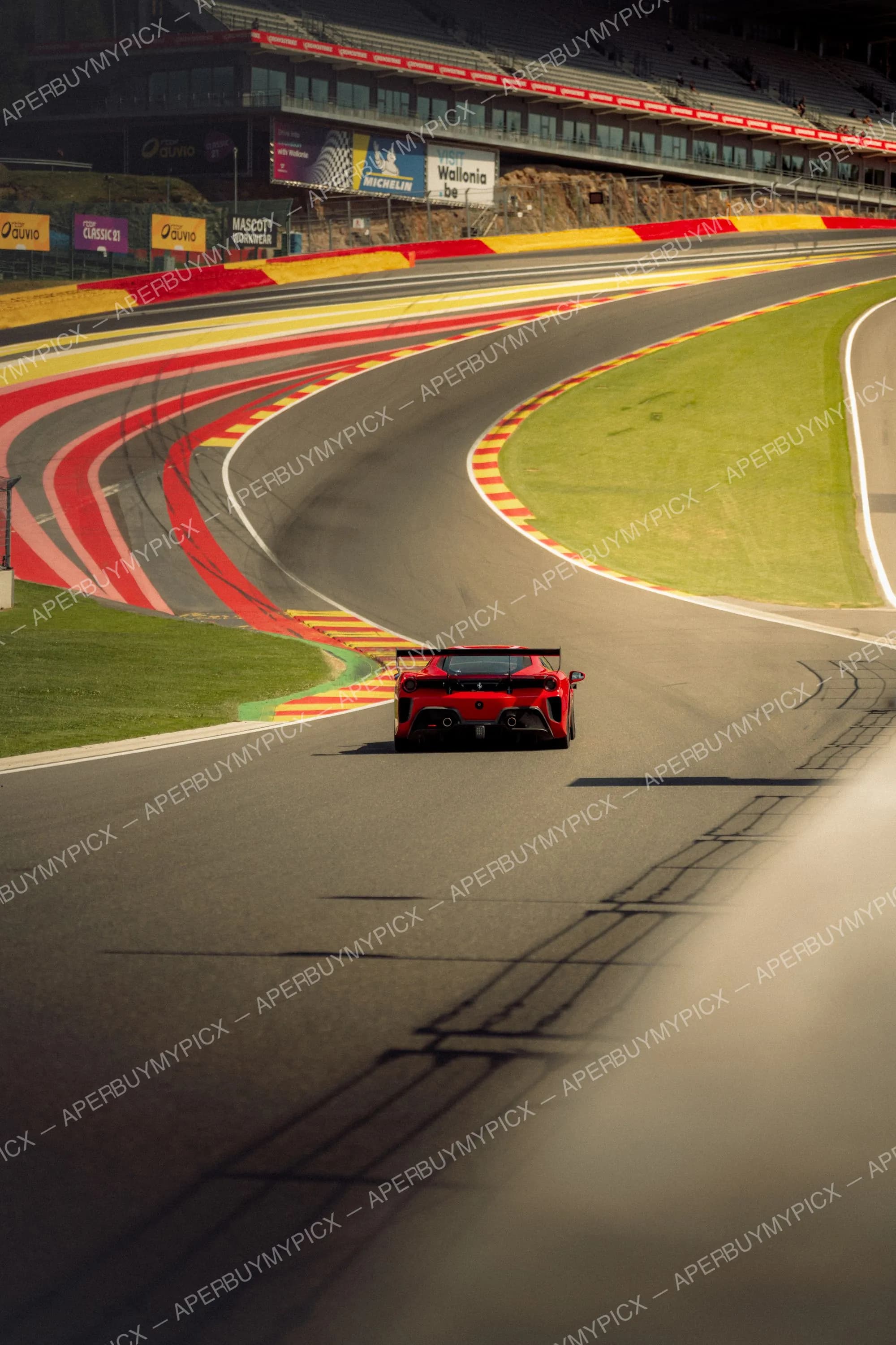 Ferrari rouge de course vue de dos filant sur la piste du circuit de Spa-Francorchamps, avec vibreurs rouges et jaunes, tribunes et panneaux Michelin et Wallonia en arrière-plan.