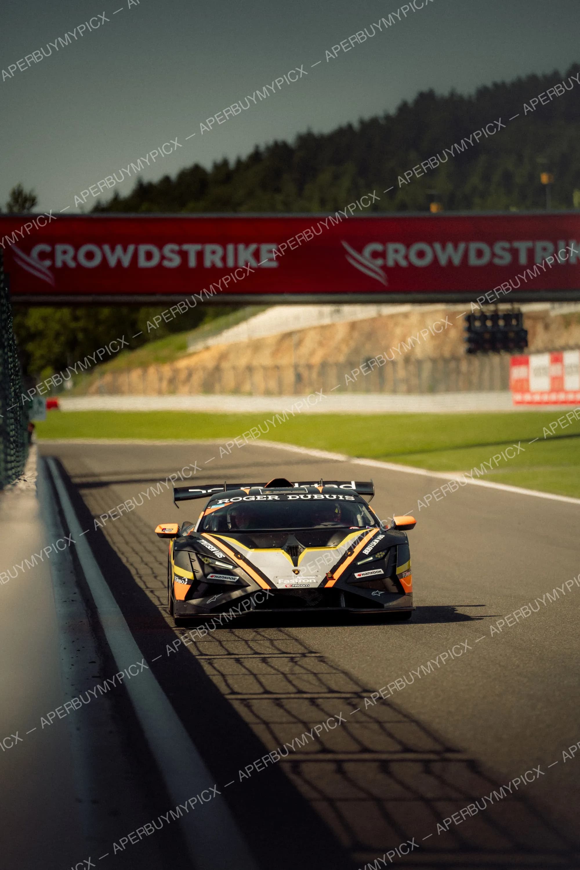 Lamborghini Huracán Super Trofeo noire, orange et jaune en piste sous une arche CrowdStrike au circuit de Spa-Francorchamps, forêt en arrière-plan.