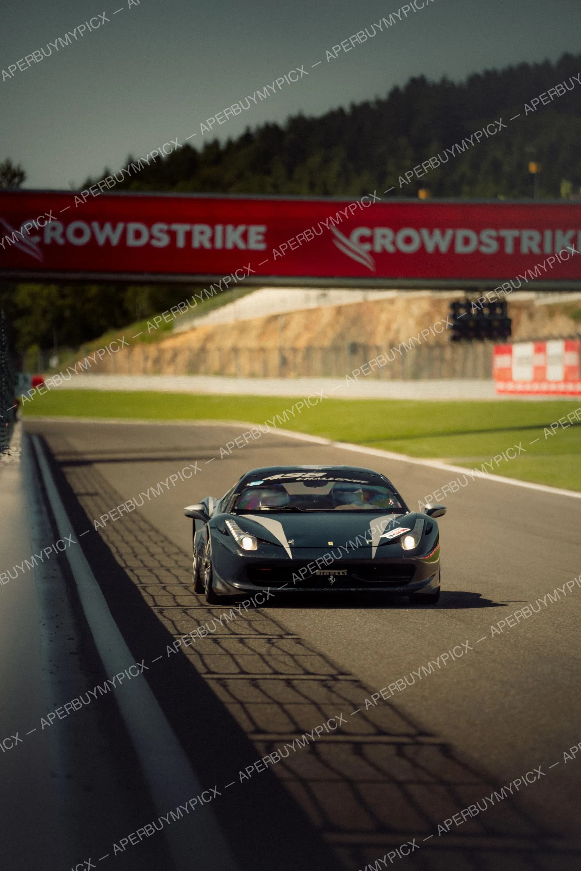 Ferrari 458 Challenge noire roulant sur la piste de Spa-Francorchamps sous un panneau publicitaire CrowdStrike rouge, entourée de collines boisées.