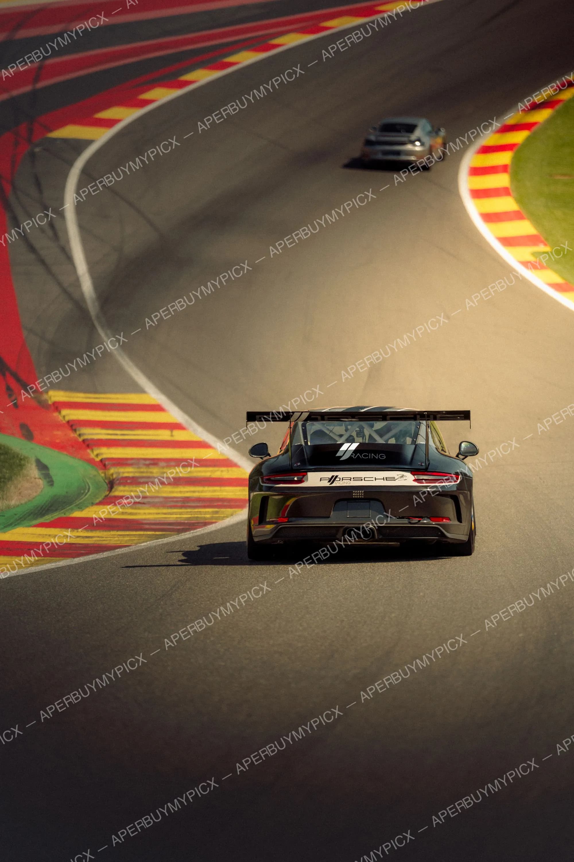 Vue arrière d'une Porsche 911 GT3 Cup noire aux couleurs Porsche Racing sur le circuit de Spa-Francorchamps, suivant une autre Porsche argentée dans un virage bordé de vibreurs rouges et jaunes.