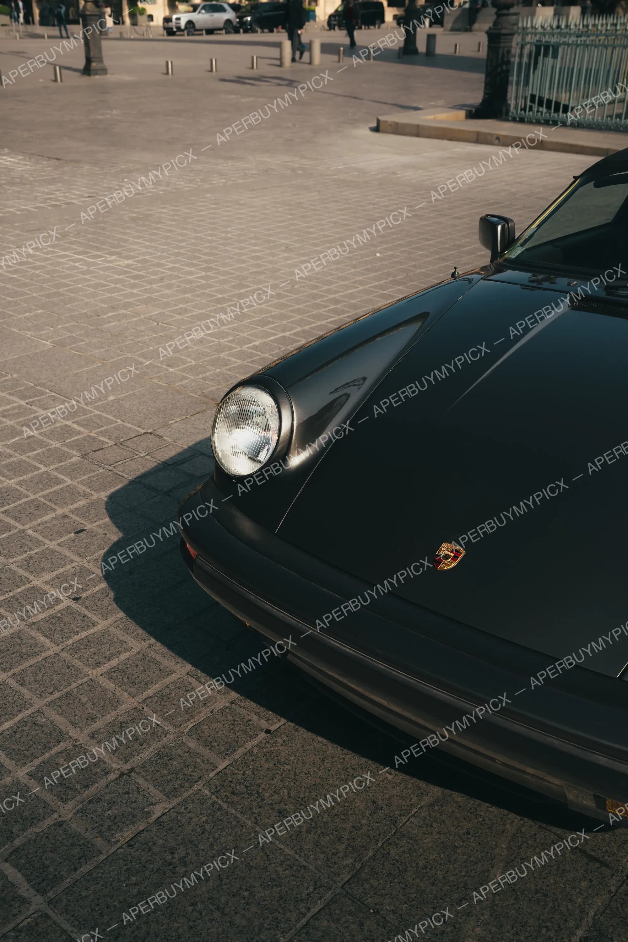 Avant d'une Porsche 911 noire vintage avec phare rond et emblème doré, garée sur pavés au coucher de soleil