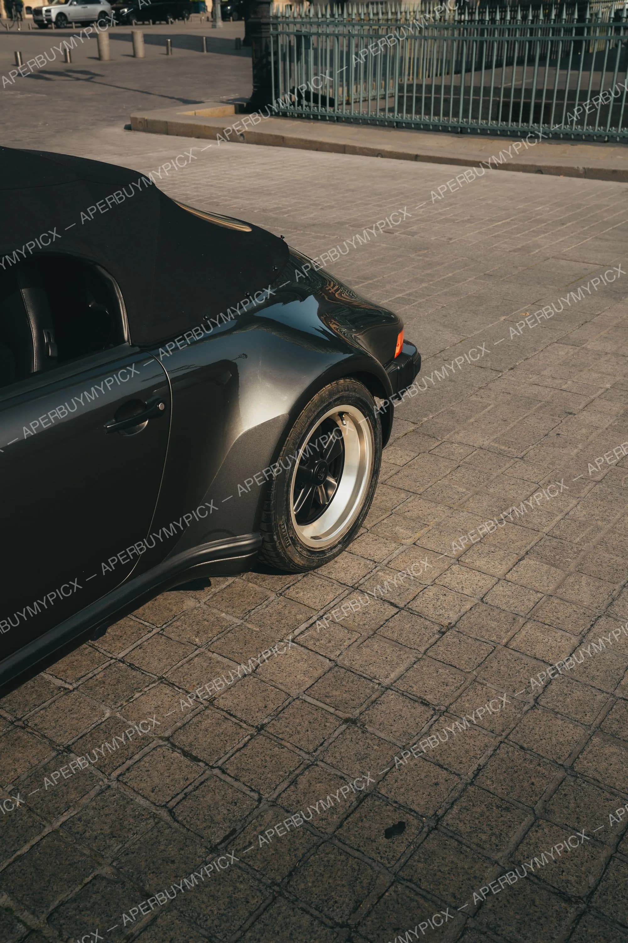 Vue arrière-latérale d'une Porsche 911 Speedster noire décapotable avec capote en toile noire et jantes Fuchs, garée sur des pavés près d'une grille métallique verte en lumière dorée.
