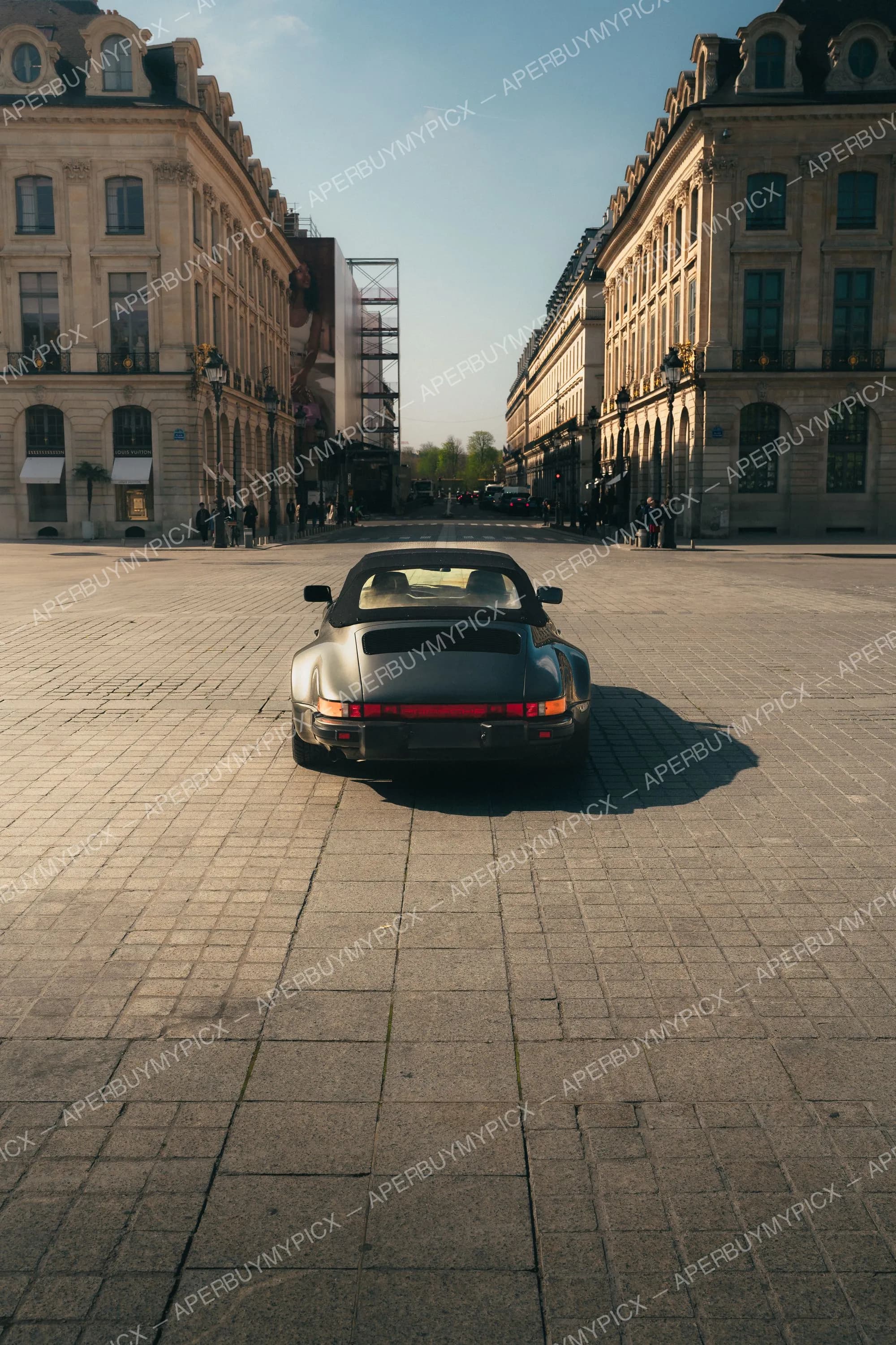 Porsche 911 cabriolet gris vue de l'arrière, stationnée sur les pavés de la Place Vendôme à Paris, entourée de bâtiments haussmanniens sous la lumière du soleil.