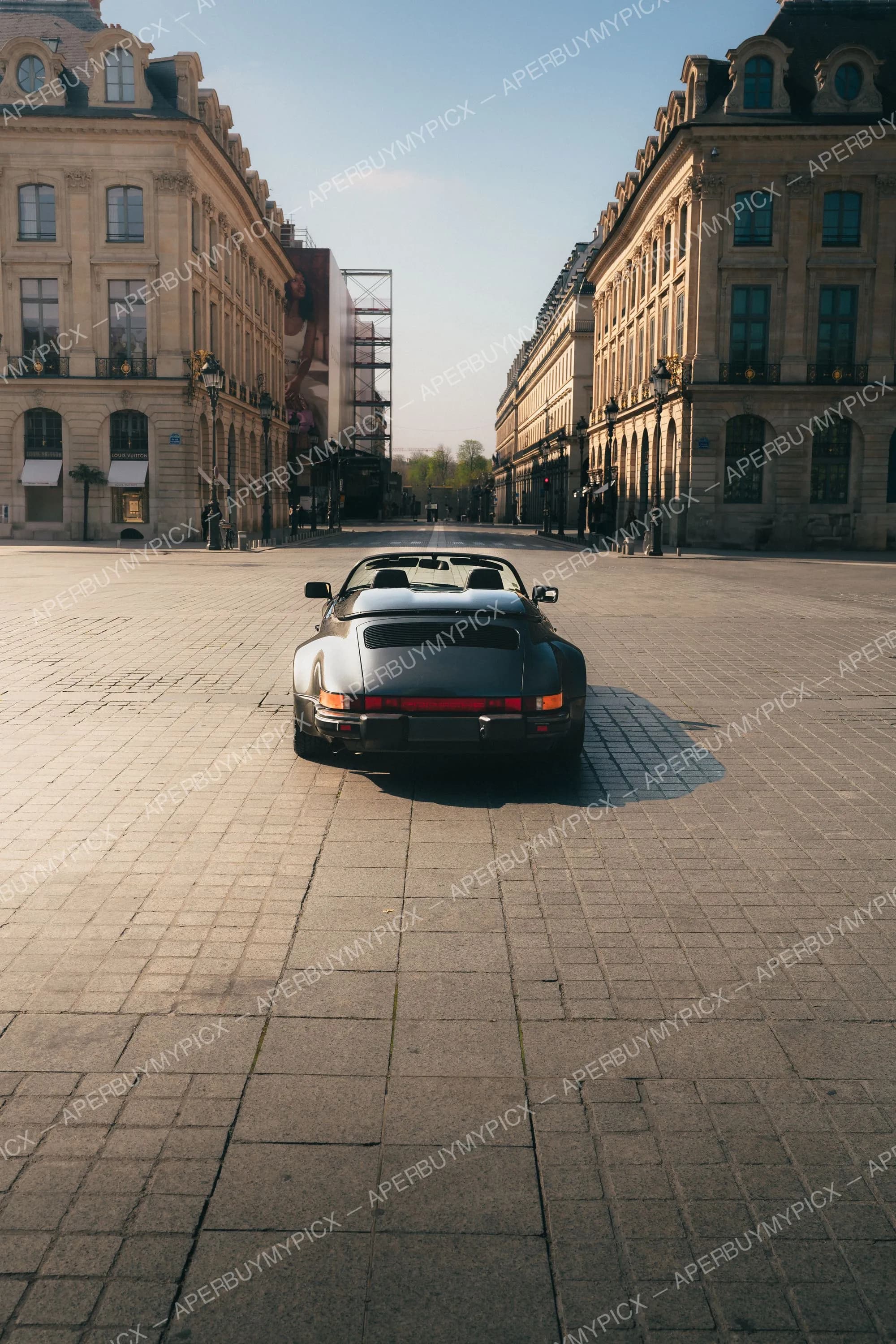 Vue arrière d'une Porsche 911 Speedster gris foncé garée sur les pavés de la place Vendôme à Paris, entourée de bâtiments haussmanniens dorés par la lumière du matin.
