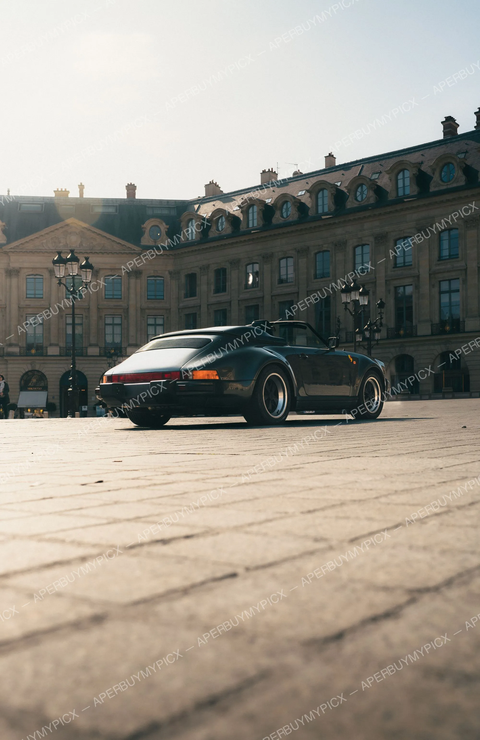 Porsche 911 Speedster noire vue de trois quarts arrière garée sur les pavés de la Place Vendôme à Paris, avec une façade haussmannienne et des lampadaires en fer forgé en arrière-plan sous une lumière dorée.