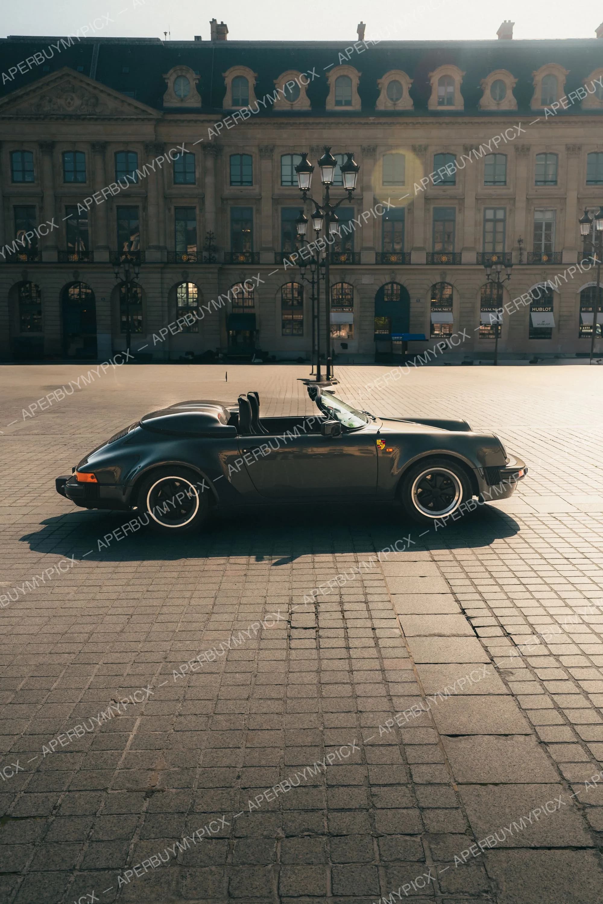 Porsche 911 Speedster gris foncé cabriolet vue de profil garée sur les pavés de la Place Vendôme à Paris, façade haussmannienne et lampadaire en arrière-plan au lever du soleil
