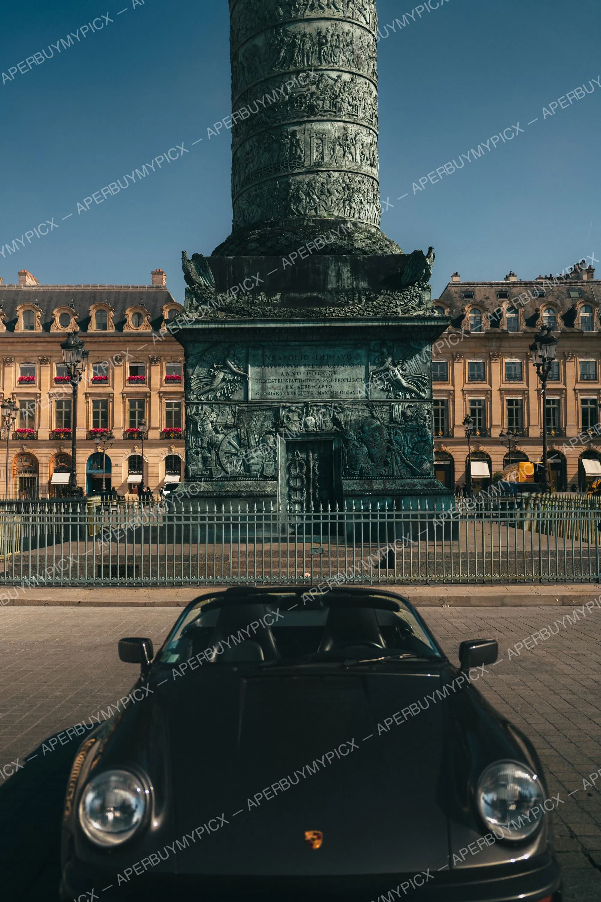 Porsche 911 cabriolet noire stationnée devant la colonne Vendôme à Paris, entourée de bâtiments haussmanniens sous un ciel bleu.