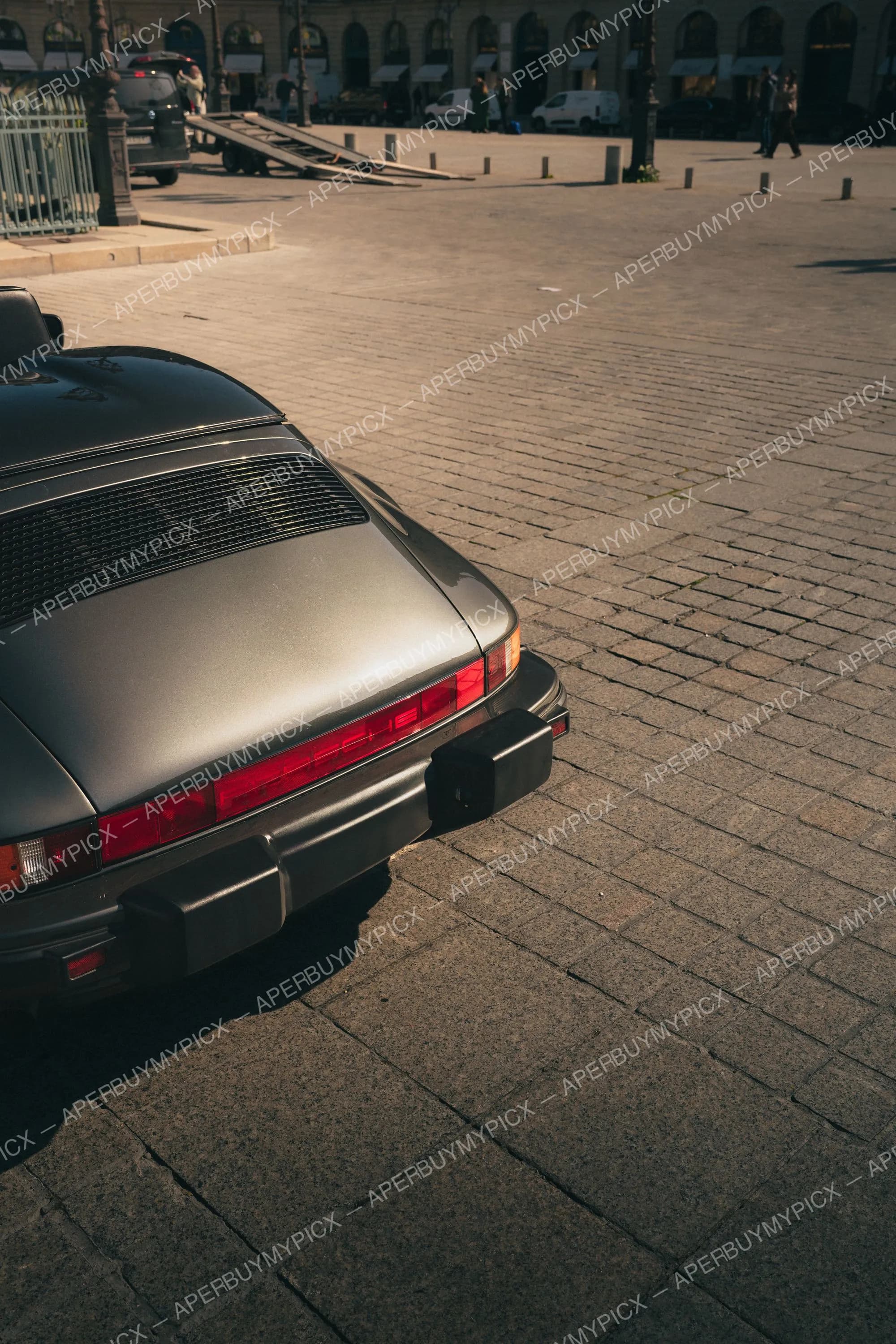 Vue arrière d'une Porsche 911 classique argentée garée sur une place pavée à Paris, avec ses feux arrière rouges éclairés par la lumière dorée du soleil, des arcades et des passants en arrière-plan.