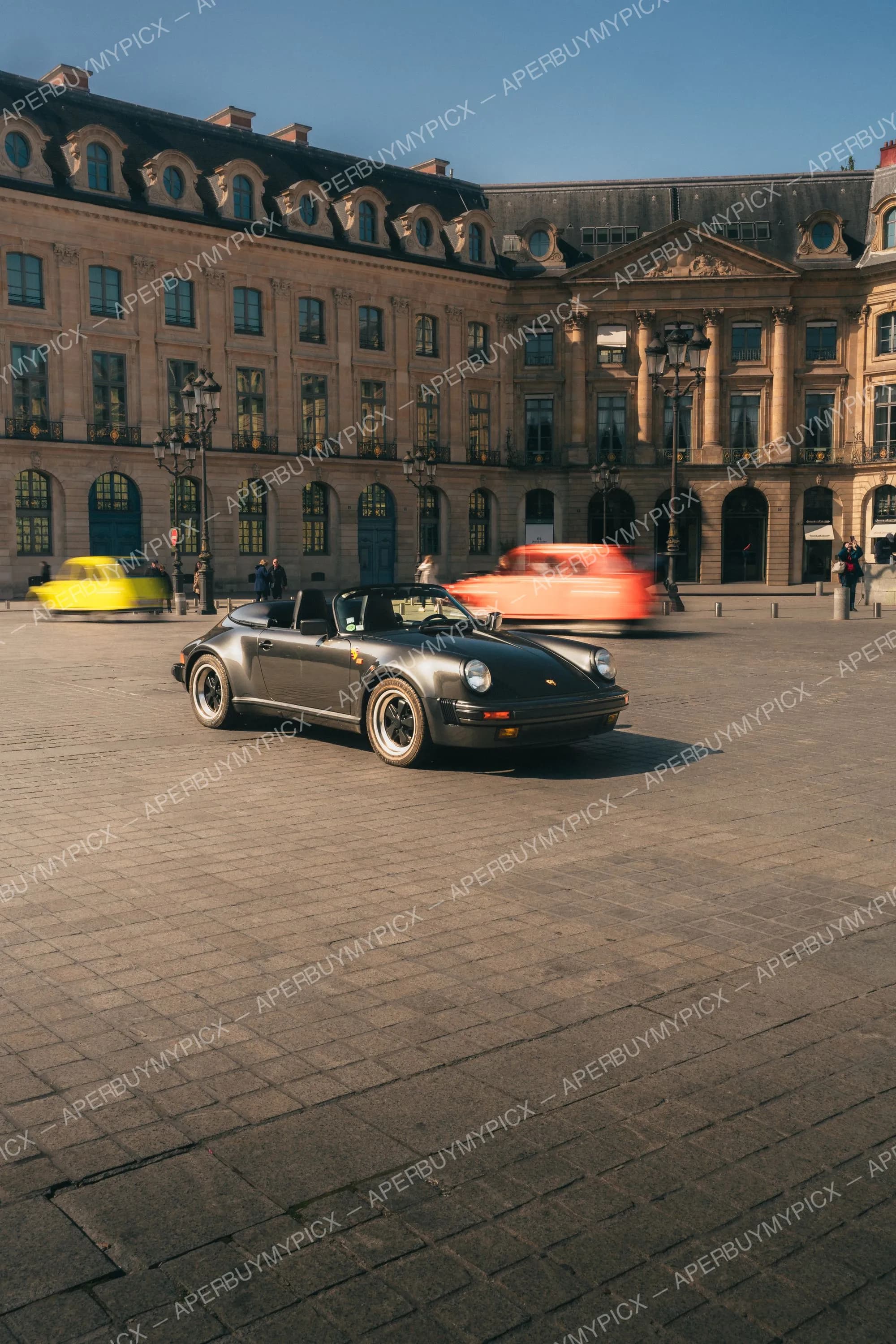Porsche 911 Speedster cabriolet gris stationnée sur les pavés de la Place Vendôme à Paris, devant un bâtiment haussmannien, avec des voitures jaune et rouge en mouvement flou à l'arrière-plan