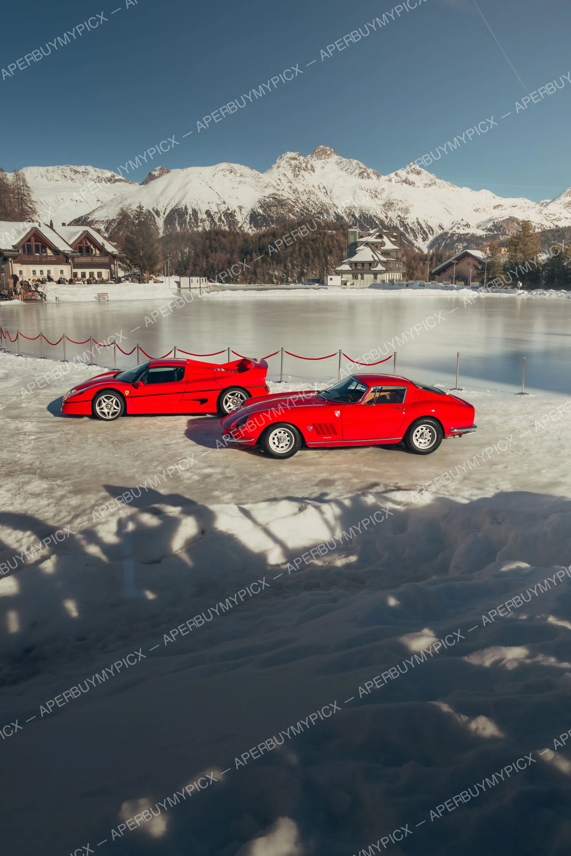 Deux Ferrari rouges, une F50 et une 275 GTB classique, garées sur un lac gelé à St. Moritz, avec chalets et montagnes alpines enneigées en arrière-plan sous un ciel bleu.