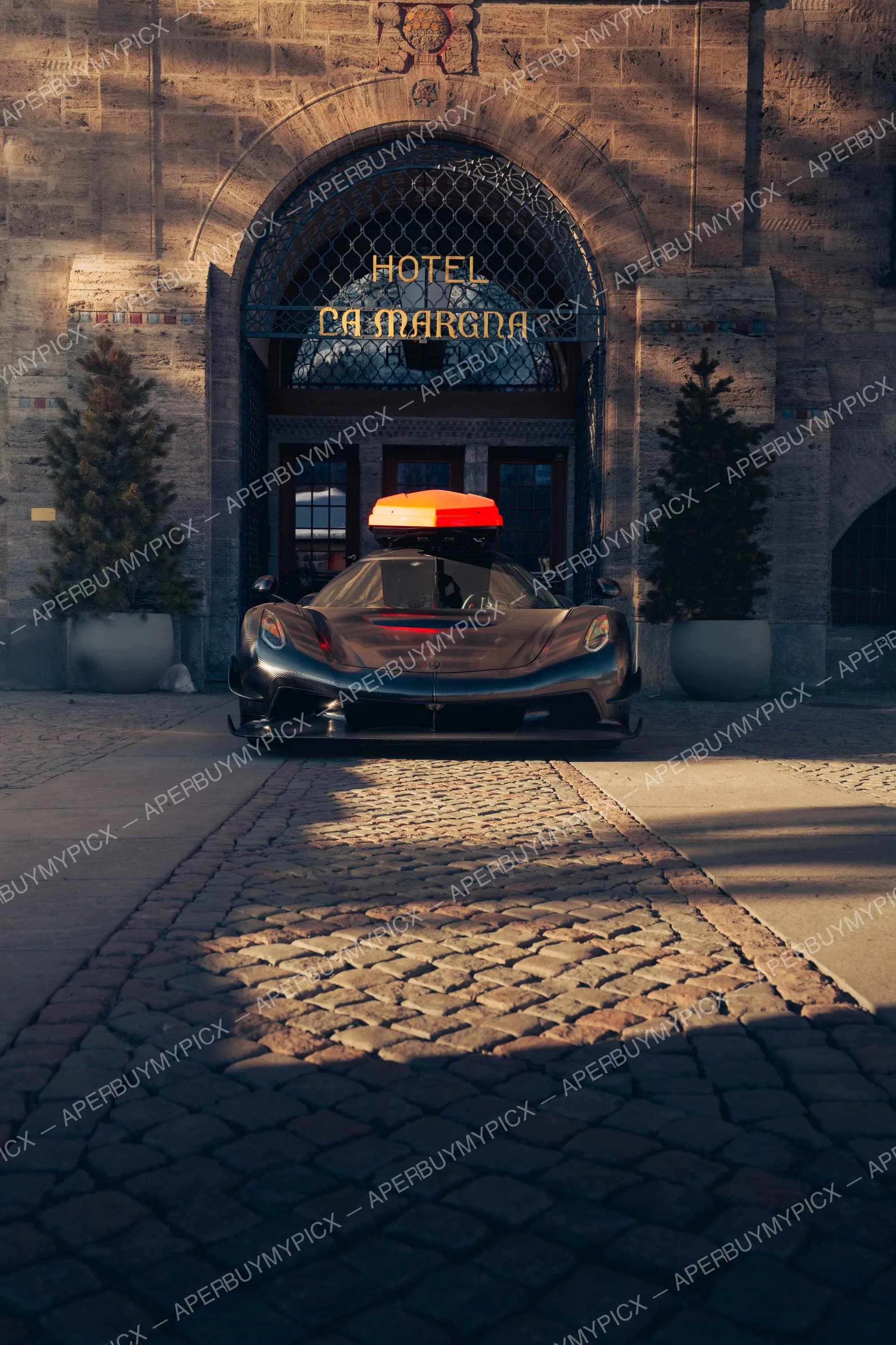 Hypercar noire en fibre de carbone avec coffre de toit orange, stationnée sur des pavés devant l'entrée arquée en pierre de l'Hôtel La Margna à St. Moritz, entourée de sapins.