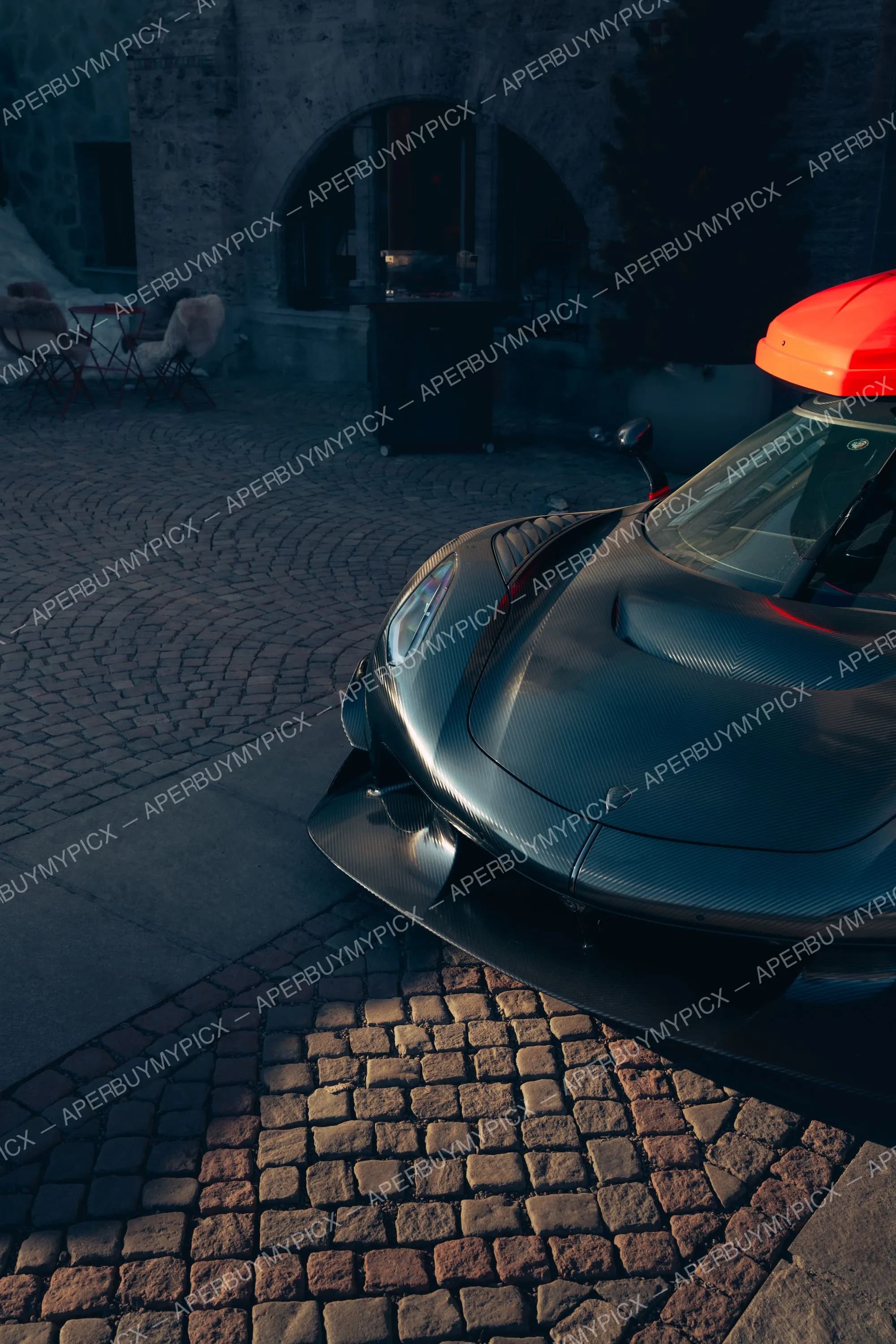 Avant d'une hypercar en fibre de carbone brute avec coffre de toit orange, stationnée sur des pavés devant un bâtiment en pierre à St. Moritz au crépuscule.