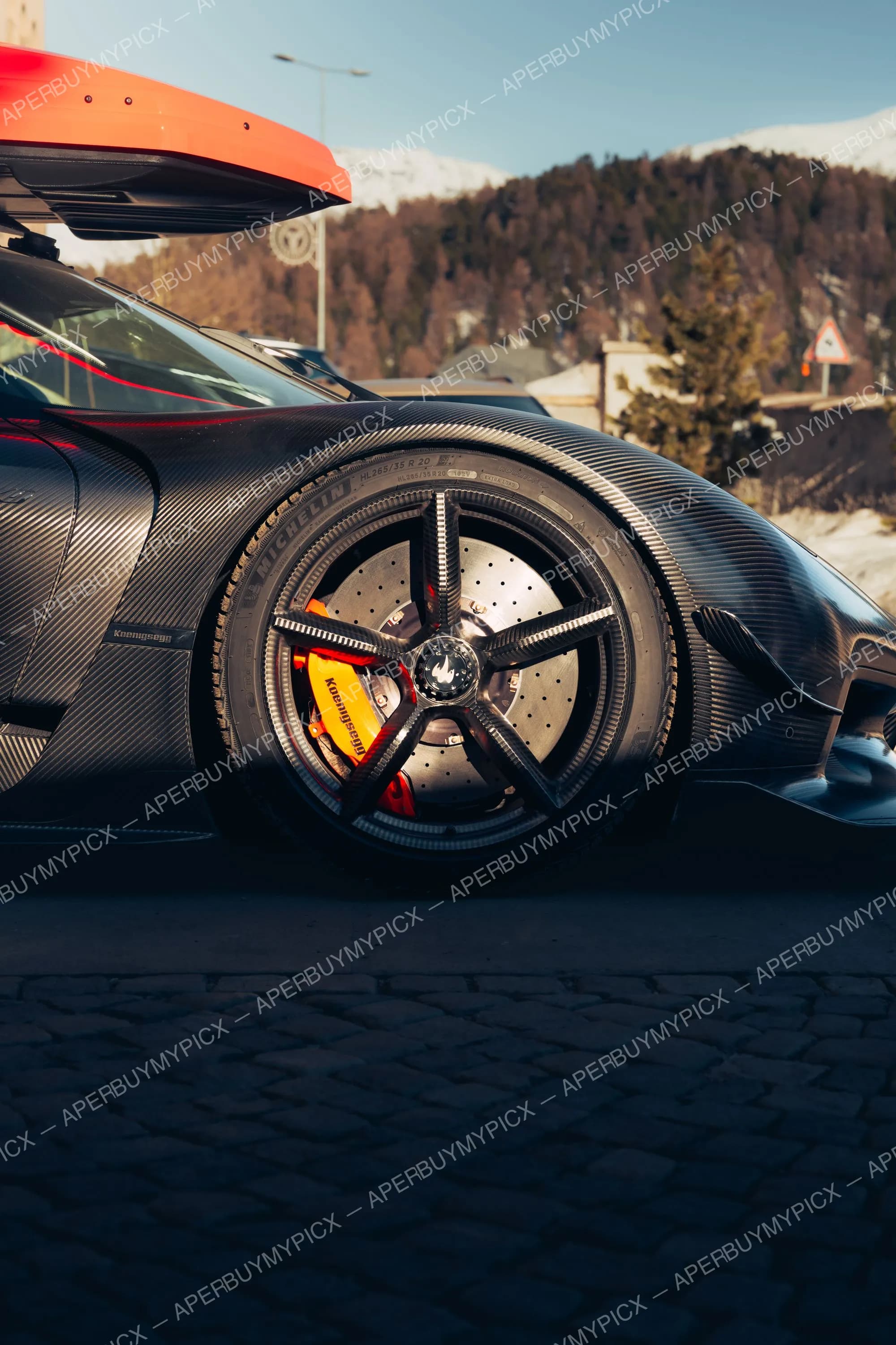 Gros plan sur la roue en carbone d'une Koenigsegg noire avec pneu Michelin, étrier de frein jaune Koenigsegg et disque céramique, sur pavés à St. Moritz avec montagnes enneigées en arrière-plan.