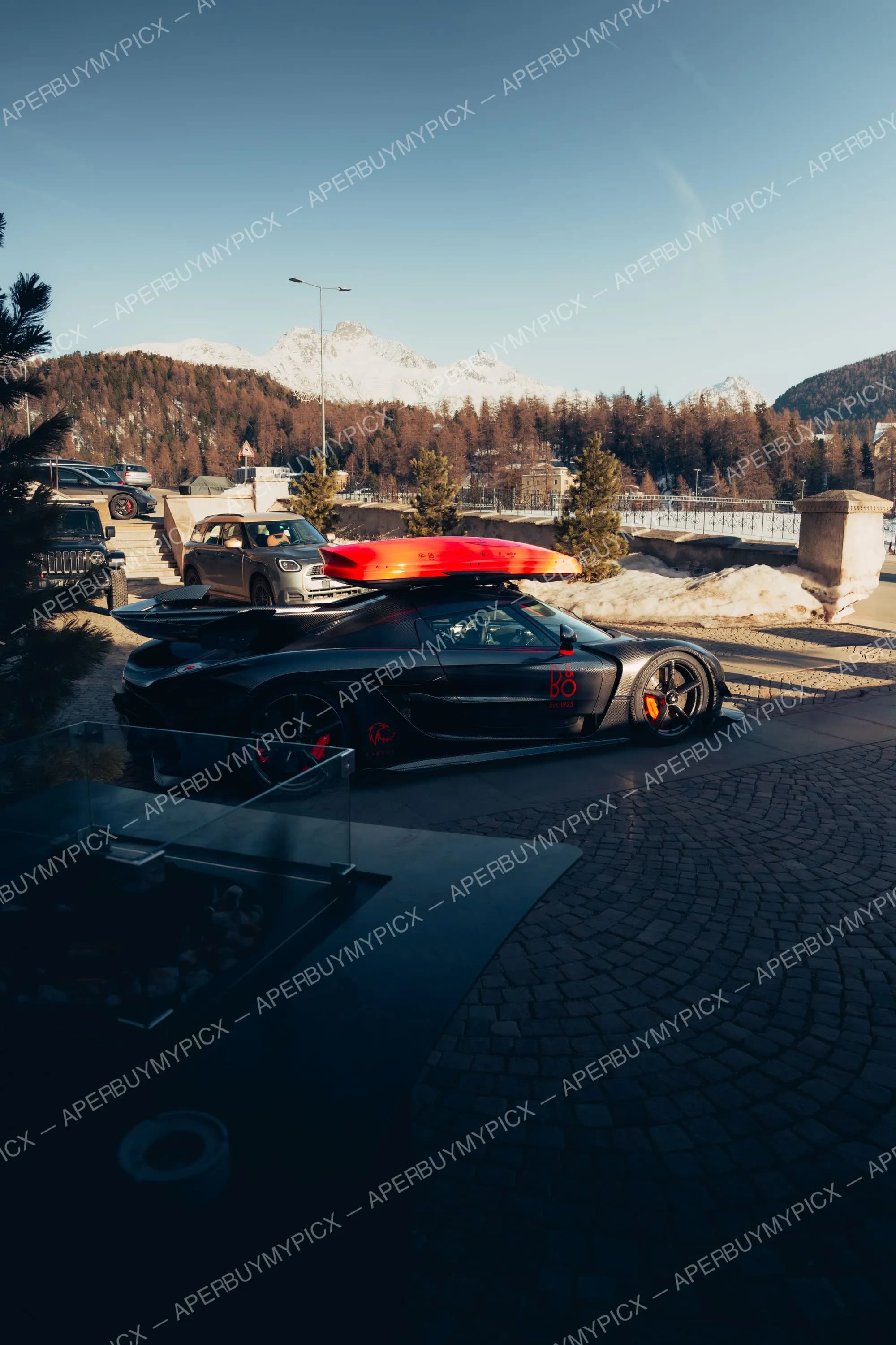 Hypercar Koenigsegg en fibre de carbone noire avec coffre de toit rouge et étriers rouges, garée sur des pavés à St. Moritz, devant des montagnes enneigées et une forêt alpine.