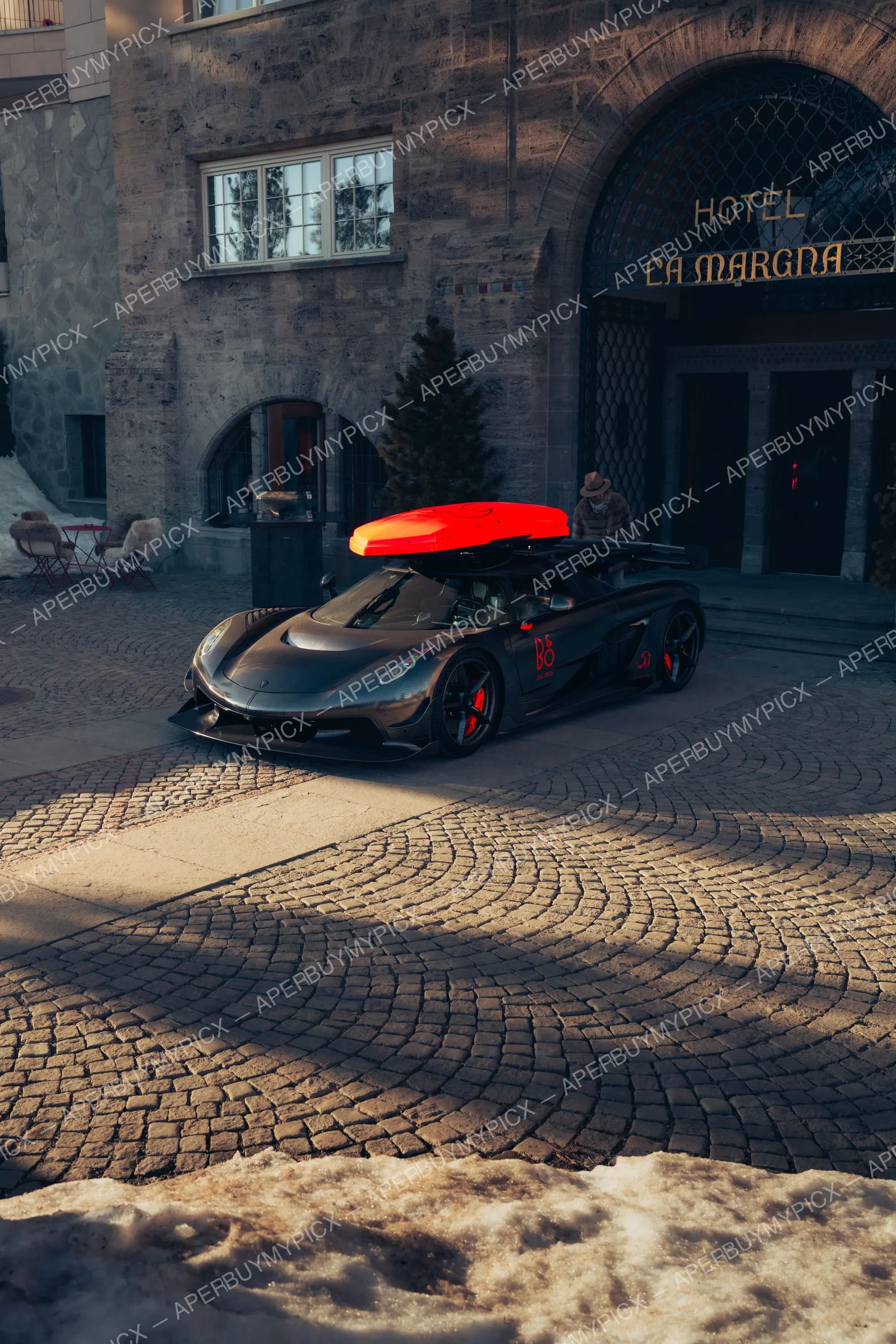 Hypercar en fibre de carbone avec un coffre de toit orange vif stationnée sur des pavés devant l'entrée en pierre de l'Hôtel La Margna à St. Moritz au coucher du soleil hivernal, neige au premier plan.