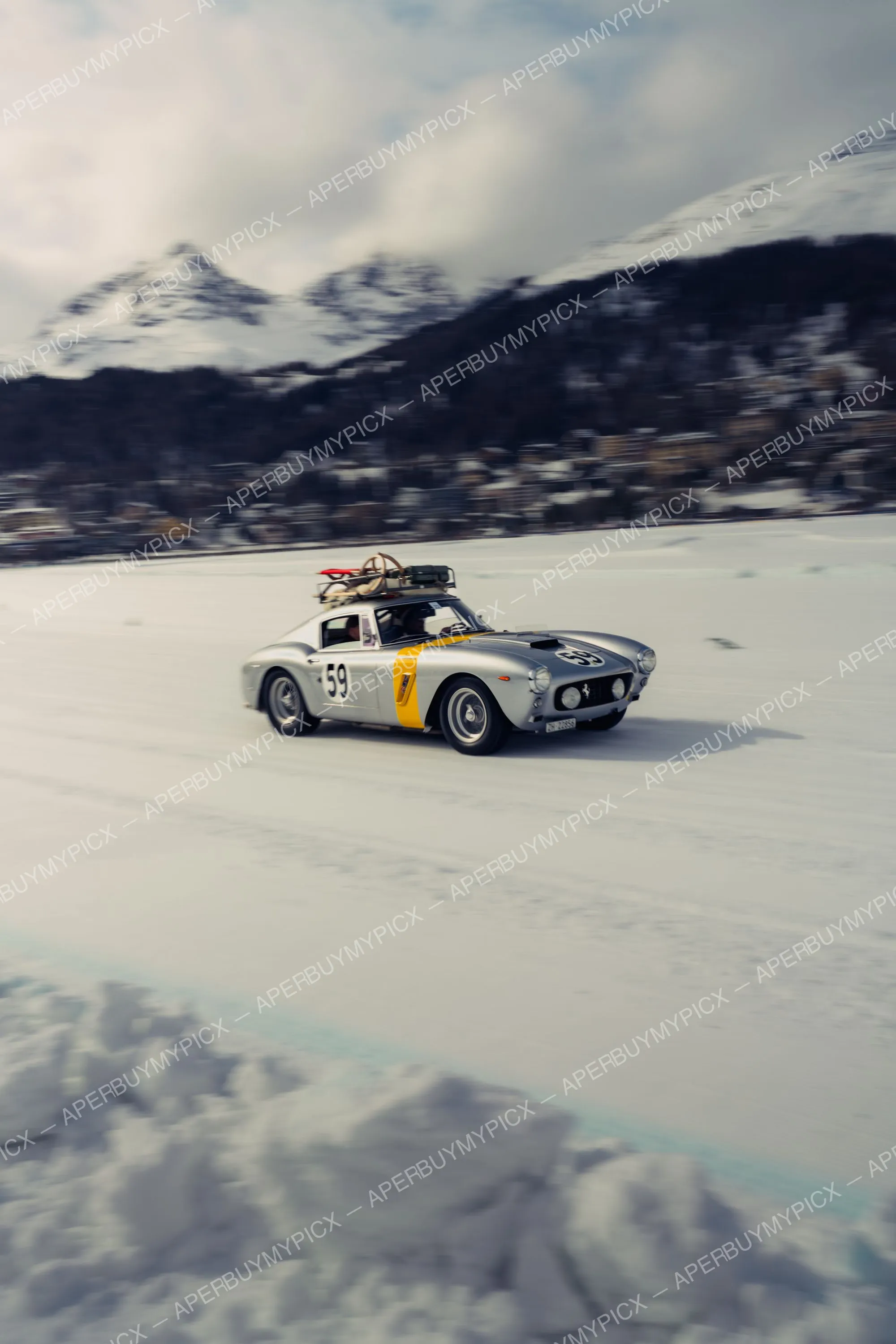 Ferrari 250 GT argentée numéro 59 avec bande jaune et skis sur le toit roulant à vive allure sur un lac gelé enneigé devant les montagnes alpines de St. Moritz