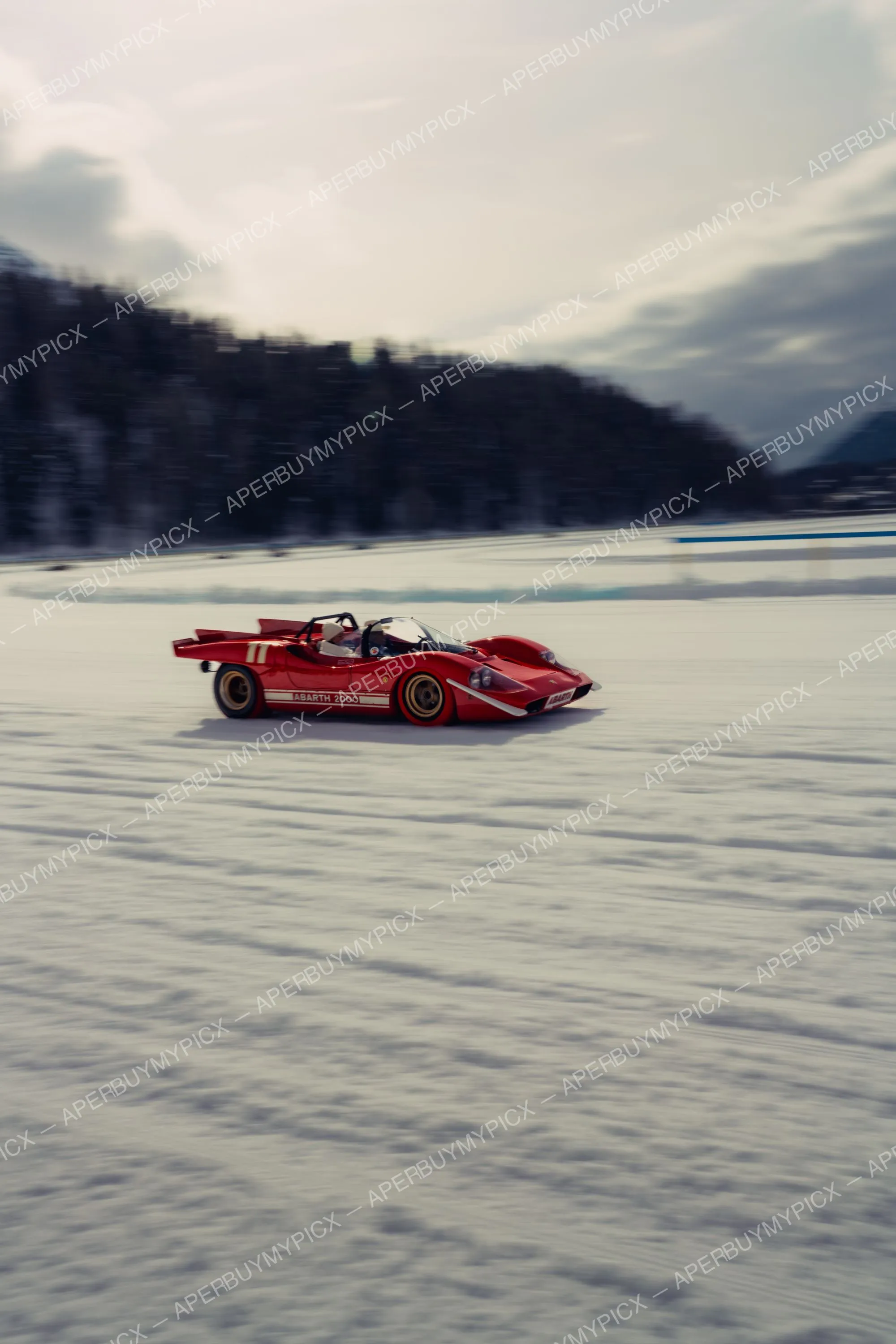 Voiture de course Abarth 2000 rouge numéro 11 filant sur un lac gelé enneigé à St. Moritz, forêt et montagnes en arrière-plan sous un ciel nuageux.