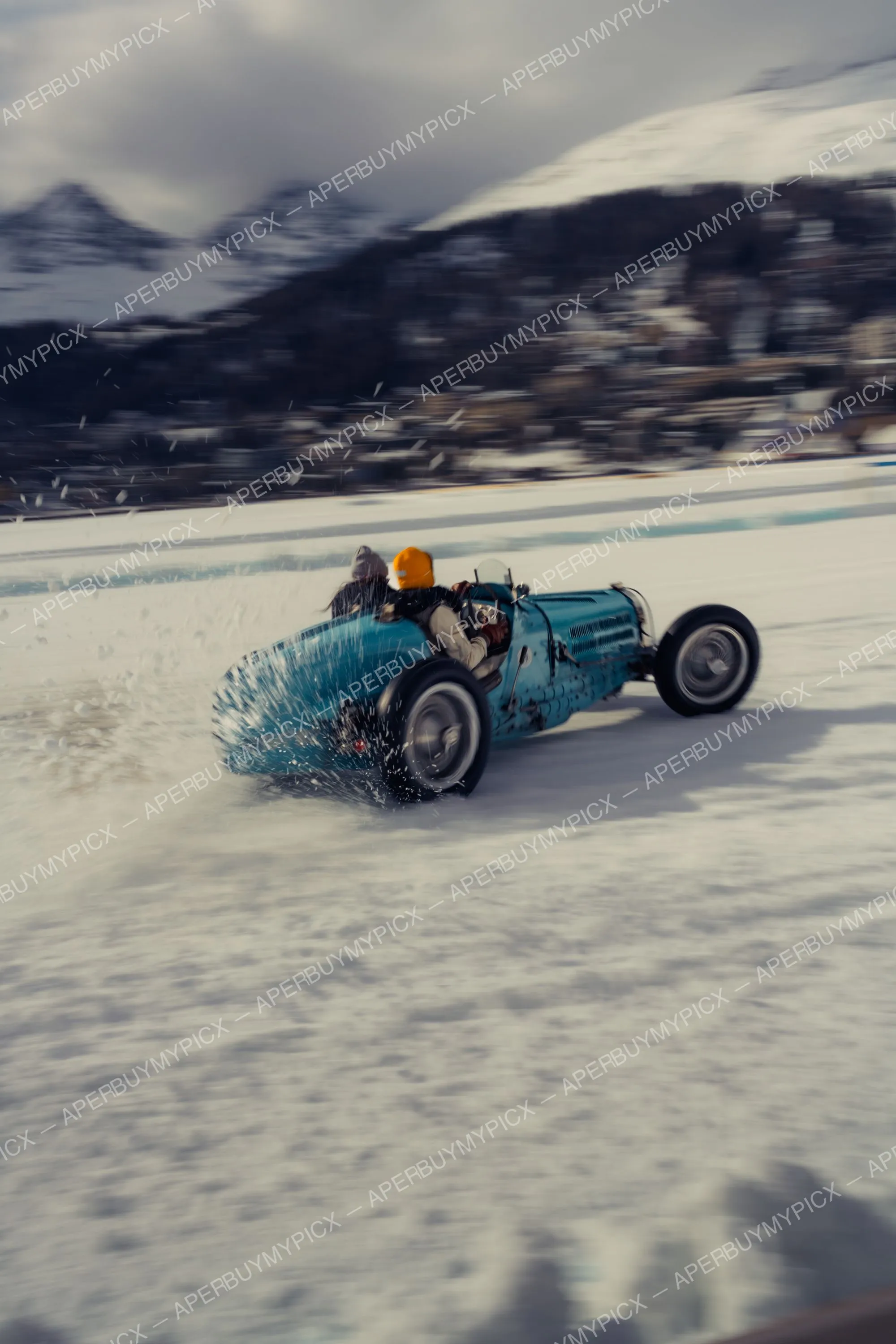 Voiture de course Bugatti bleue vintage projetant de la neige lors d'une course sur glace à St. Moritz, avec pilote au bonnet orange et montagnes enneigées en arrière-plan
