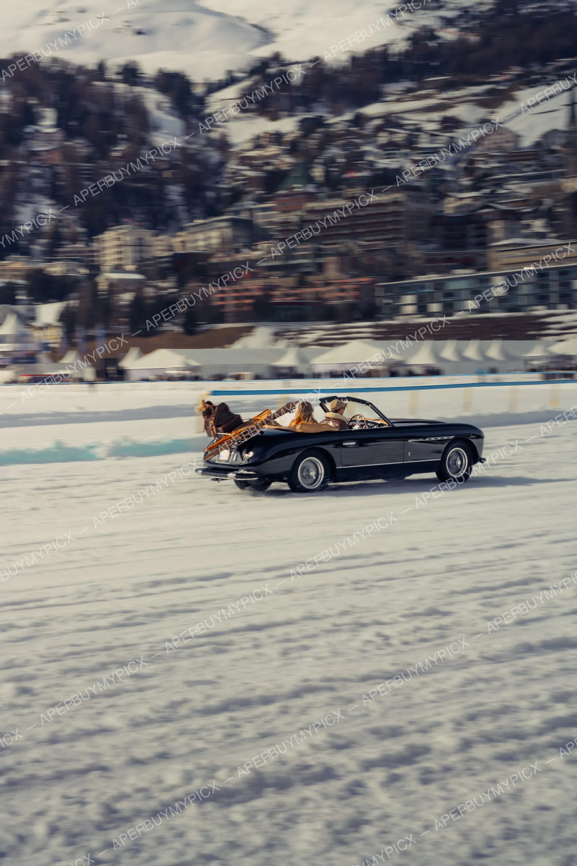 Cabriolet vintage noir roulant à vive allure sur un lac gelé à St. Moritz, avec des passagers et un traîneau à cheval en arrière-plan, montagnes enneigées derrière