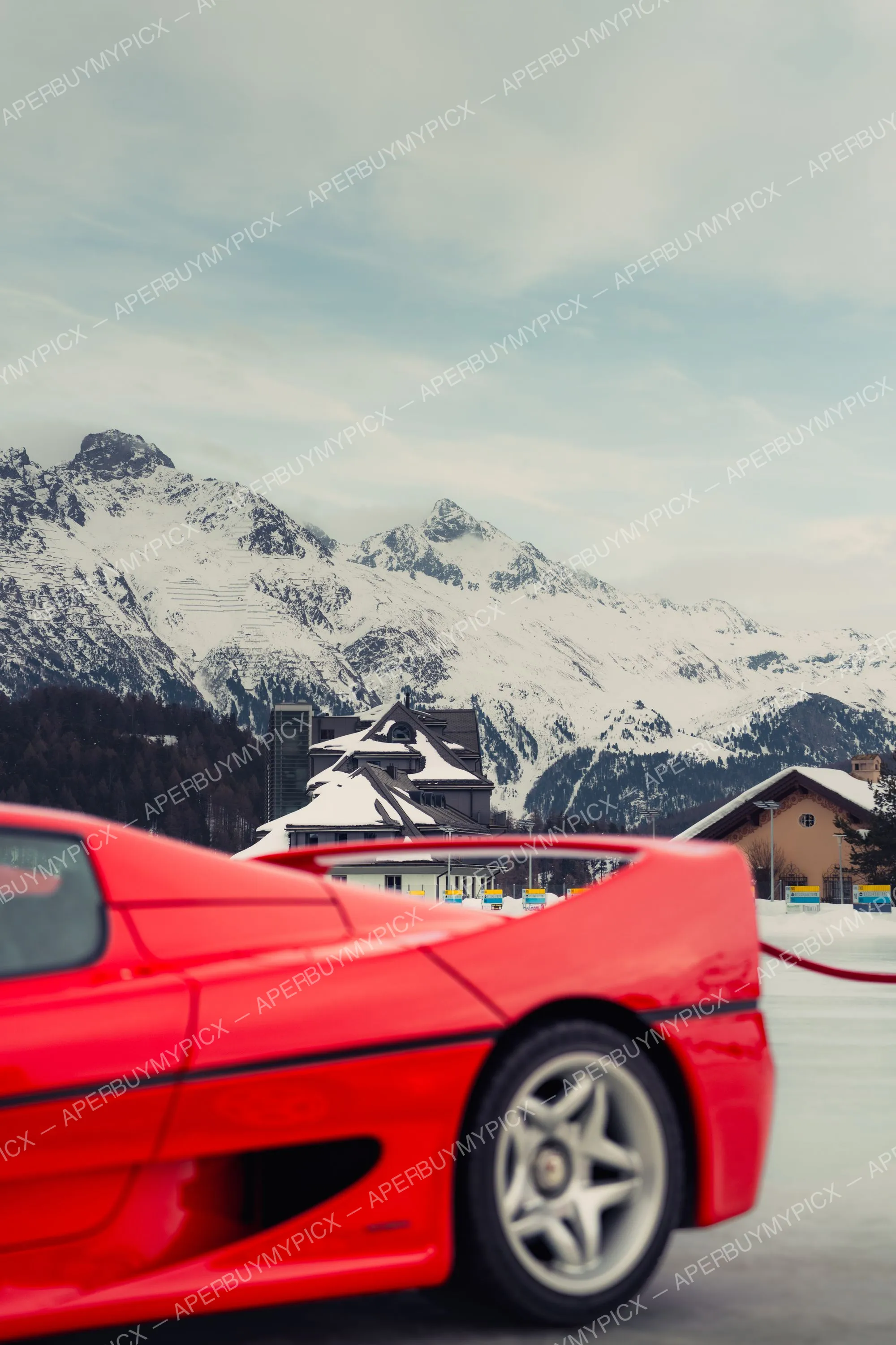 Détail arrière d'une supercar Ferrari rouge avec aileron et jante étoile, garée devant un chalet alpin et des montagnes enneigées à St. Moritz sous un ciel nuageux.