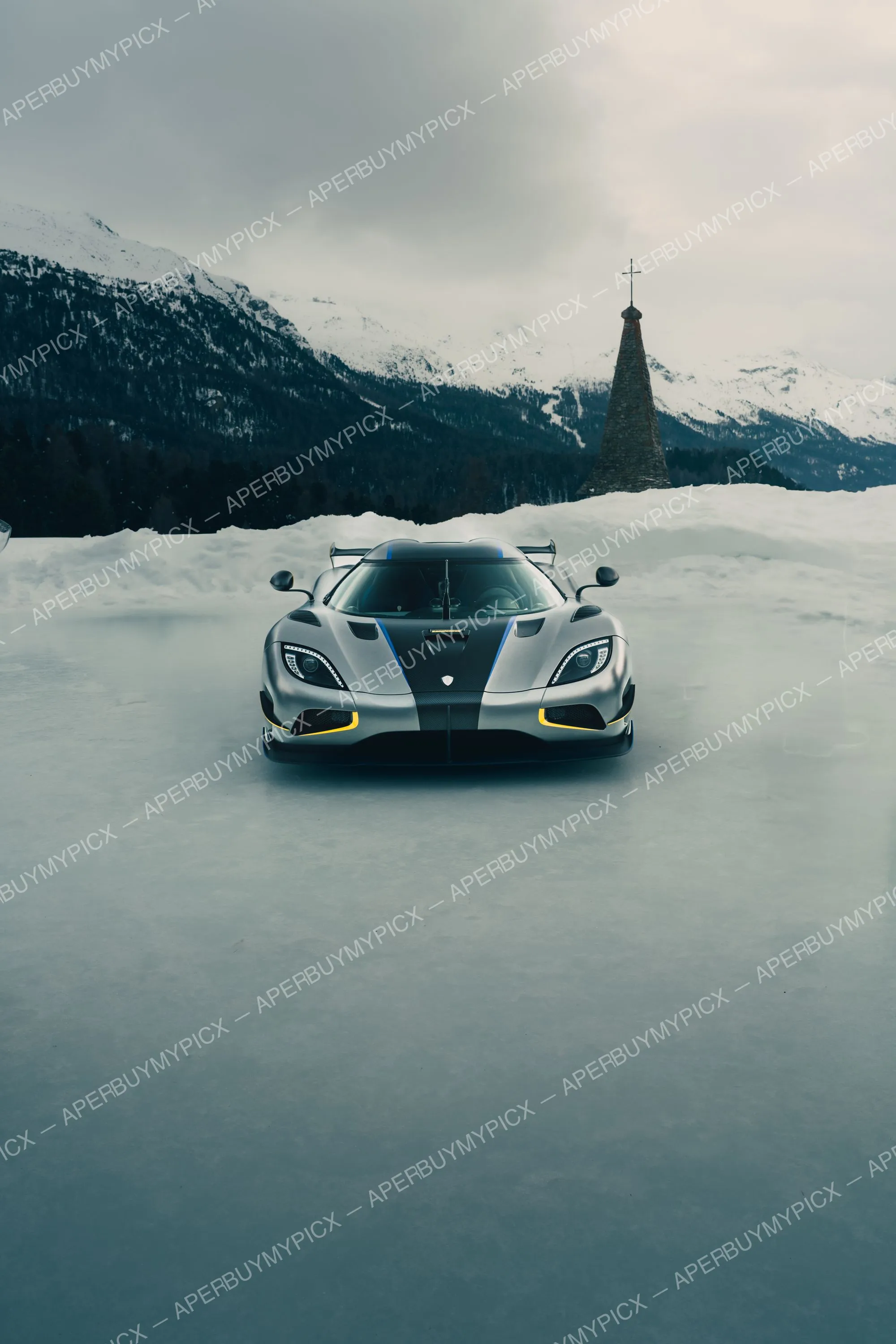 Supercar Koenigsegg grise avec détails jaunes et bleus, garée sur un lac gelé, montagnes enneigées et clocher d'église en arrière-plan sous un ciel gris à St. Moritz