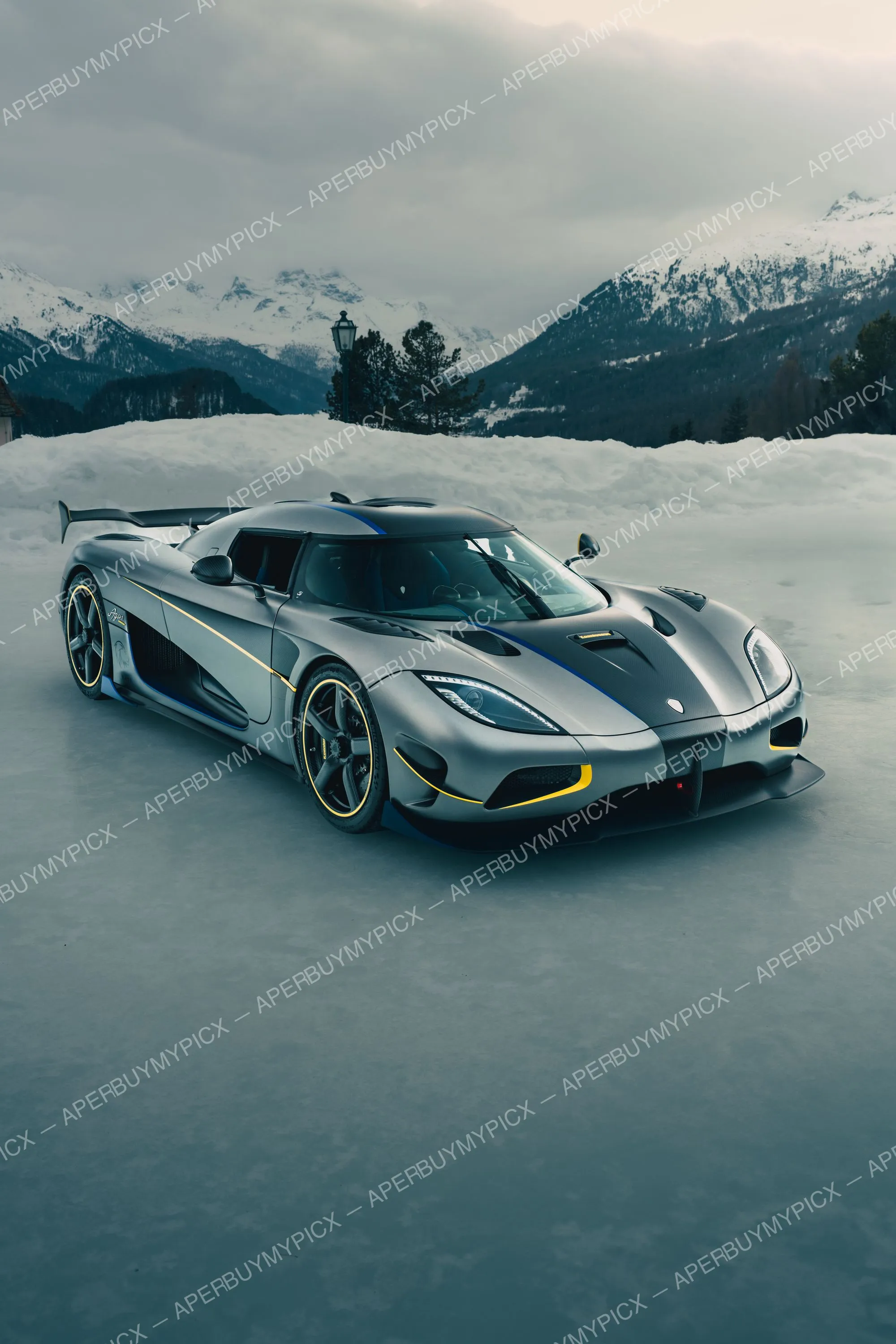 Hypercar Koenigsegg Agera grise à liseré jaune et bleu, garée sur une surface glacée devant des montagnes enneigées et une forêt sous un ciel gris à St. Moritz.