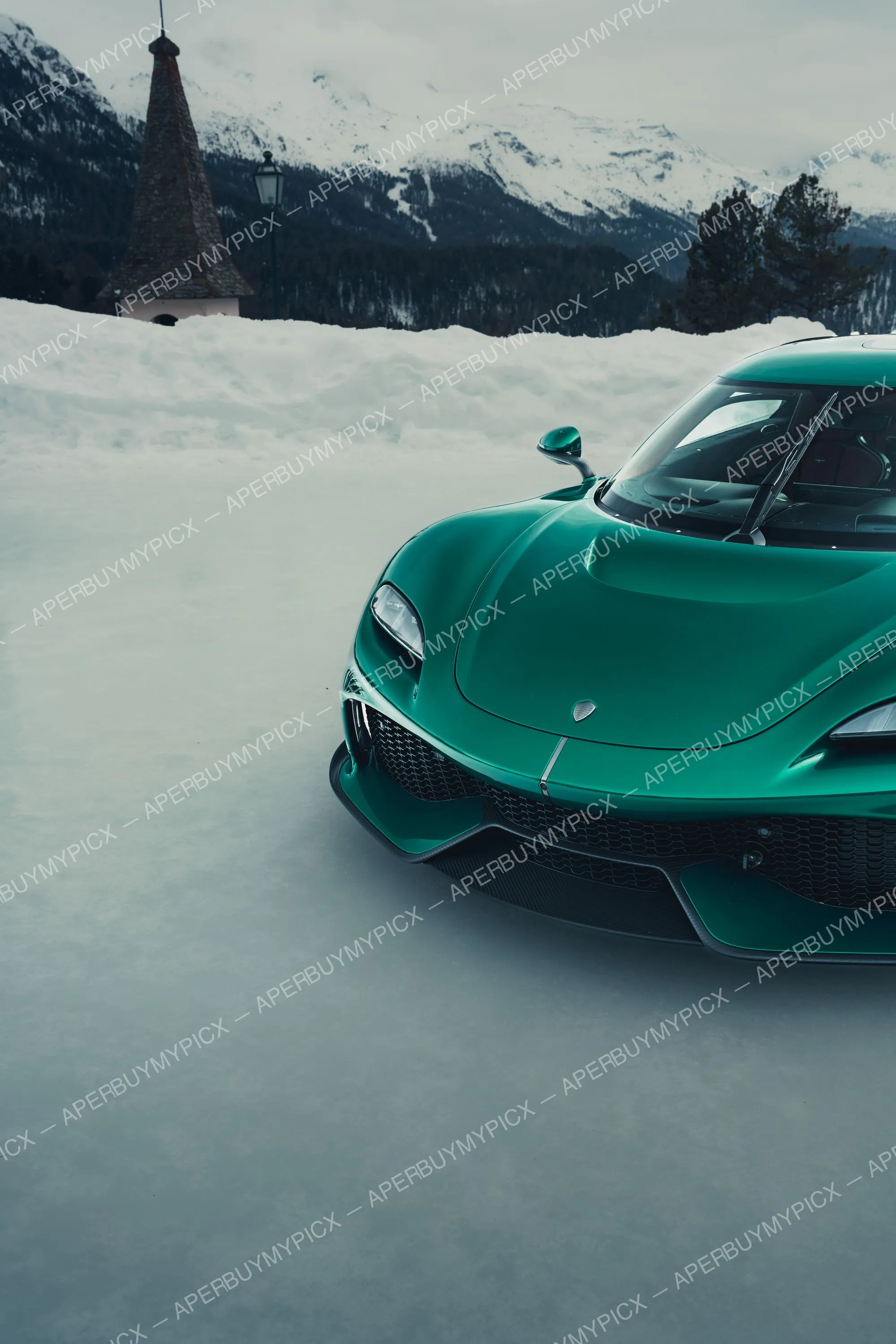 Hypercar verte garée dans la neige à St. Moritz, avec montagnes enneigées, forêt de sapins et clocher en arrière-plan sous un ciel gris d'hiver.